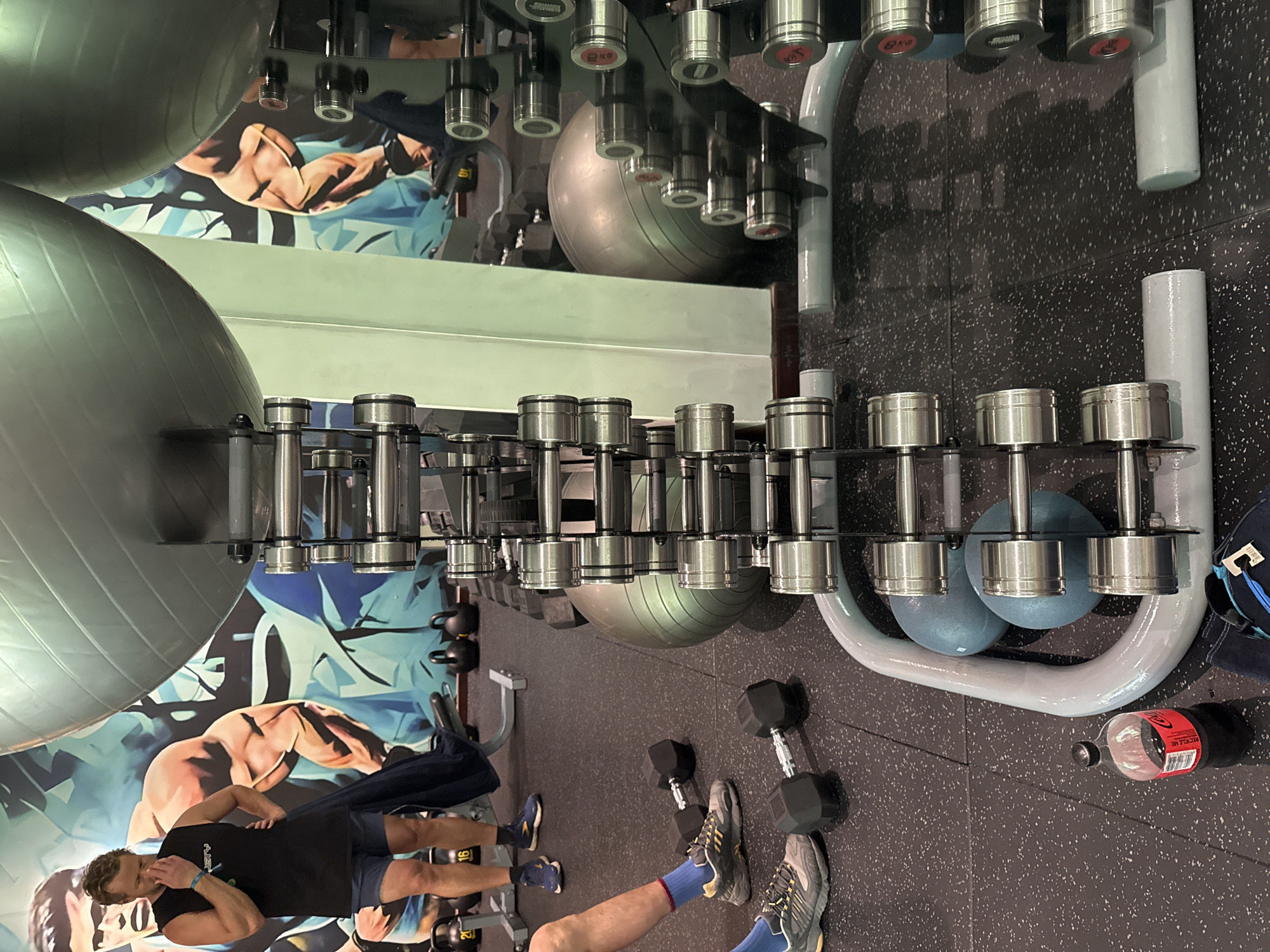 In a gym located in Kuta Selatan, Indonesia, a man is standing near a rack of dumbbells, touching his face with his hand, appearing to be in thought or discomfort. He is wearing a black sleeveless shirt and blue shorts. In the background, a large mural depicts muscular figures. Dumbbells of various weights are arranged on racks, and exercise balls are also present. On the floor, several pairs of dumbbells and a bottle of Coca-Cola are visible. The gym floor is black with speckles.
