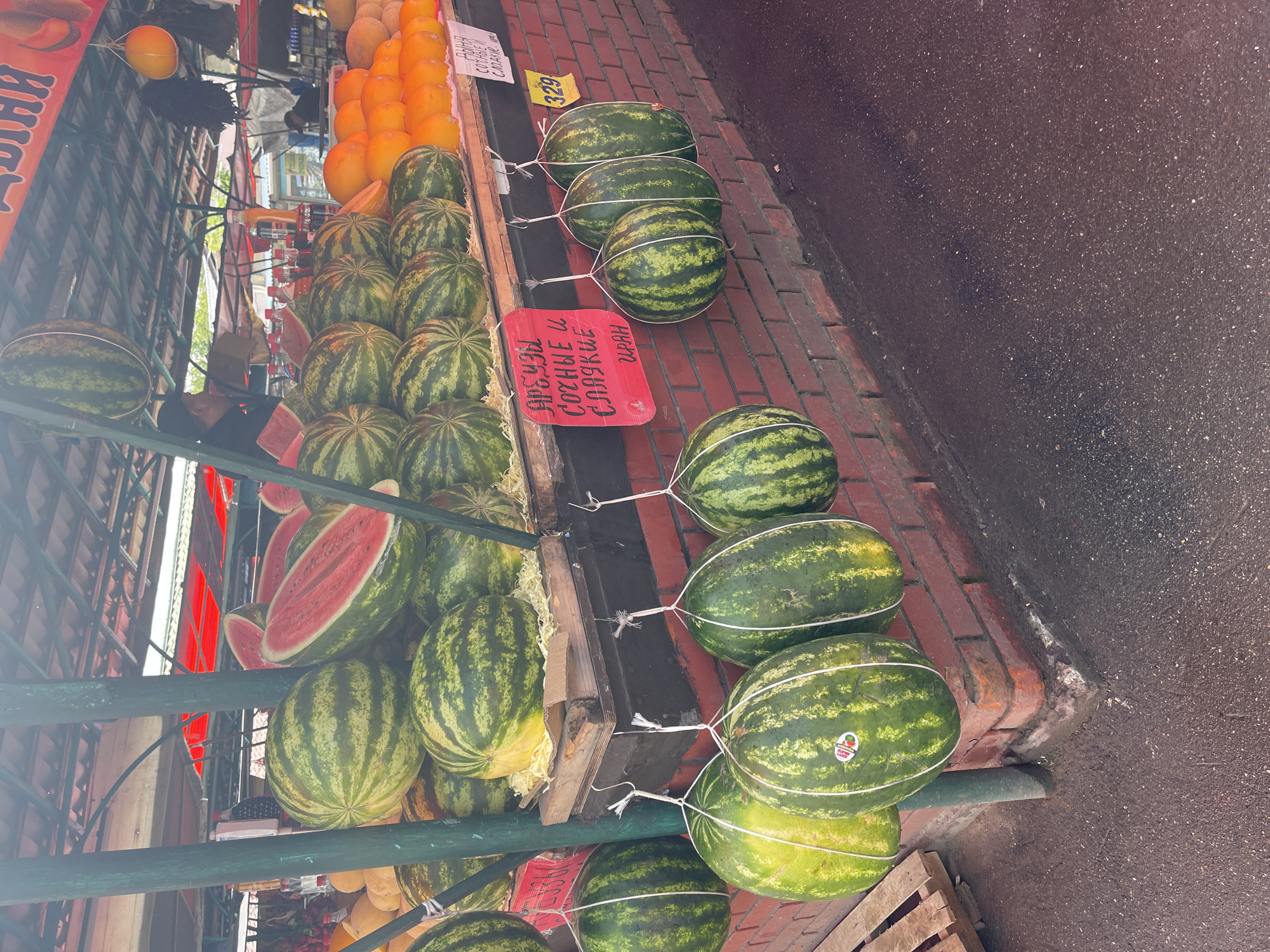 The image shows a fruit stall in what appears to be a street market. Several watermelons are displayed, some whole and tied with string, others cut open to show the red flesh. Oranges or melons are also visible in the background. A sign with writing in a Cyrillic script is present. The location is Unknown, Unknown. The pavement and street surface appear slightly wet.