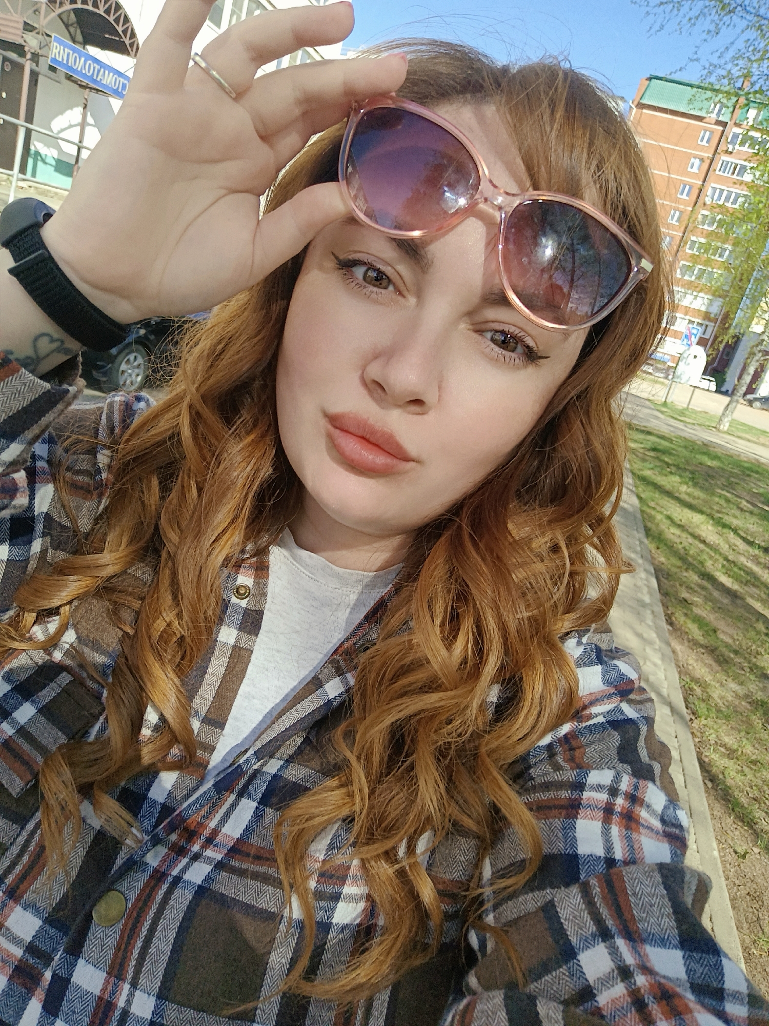 A young woman with auburn curly hair, wearing a plaid shirt and a light-colored t-shirt, holds pink sunglasses in her left hand, partially obscuring her forehead. She is outdoors, likely posing for a selfie. A black smartwatch is visible on her left wrist.
In the background, an urban environment is discernible, with an apartment building and street signs indicating the location as the city of Chaikovsky, Russia. A car is parked in the mid-ground, further suggesting an urban setting. Trees are also present, suggesting a park or green space. The time of day appears to be daytime due to the presence of natural lighting.