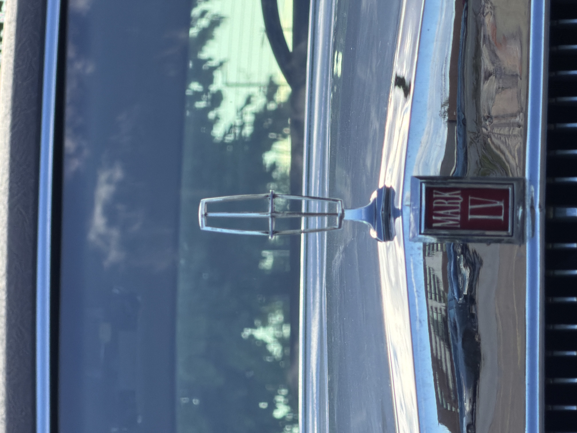 The image shows a close-up of a Lincoln Mark IV hood ornament and grille. The ornament is centered in the frame, and the "MARK IV" emblem is visible. Reflections of buildings and trees are visible on the vehicle's surface. The image was taken in Morton Grove, United States of America.