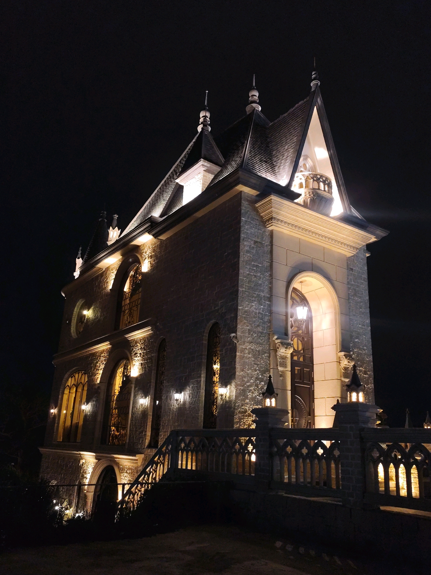 The image shows a night view of a stone building with Gothic architectural elements in Da Lat, Vietnam. The building is illuminated by warm lights, highlighting its textured stone facade, arched windows with decorative grilles, and a steeply pitched roof topped with spires. A stone balustrade with intricate patterns and decorative lanterns lines a walkway leading towards the entrance. The overall impression is one of an old-world, possibly historical or themed, structure set against a dark night sky.