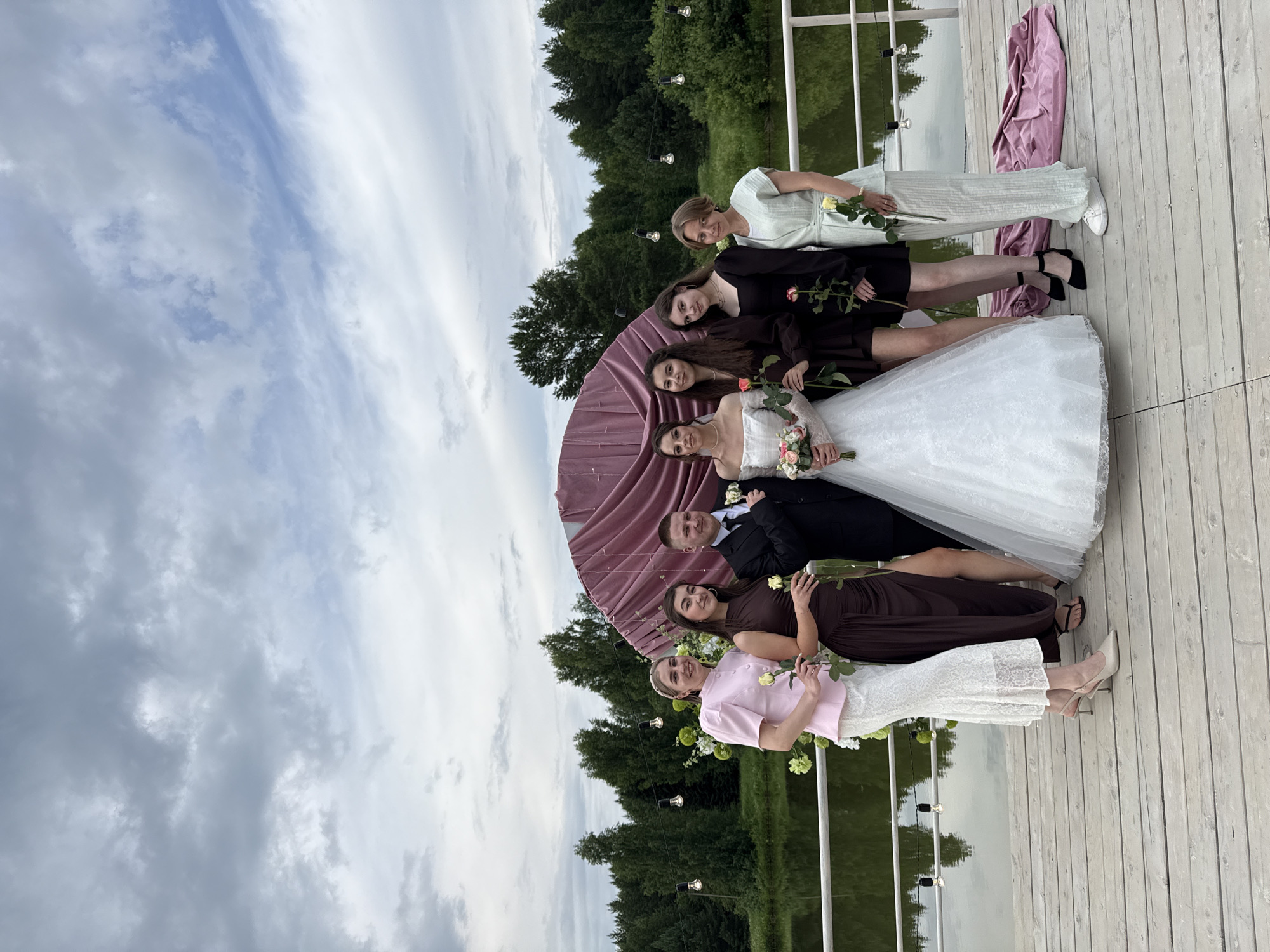 In Votkinsk, Russia, on a wooden pier overlooking a body of water, a wedding party of seven people is posing for a photograph. The group includes a bride in a white wedding dress and a groom in a dark suit, both holding roses. They are flanked by five women, likely bridesmaids, dressed in various colored gowns and holding roses as well. A decorative backdrop of dark pink fabric and floral arrangements is visible behind them. The environment suggests an outdoor wedding celebration, with trees lining the water and a cloudy sky overhead. The pier itself is constructed from light-colored wooden planks.