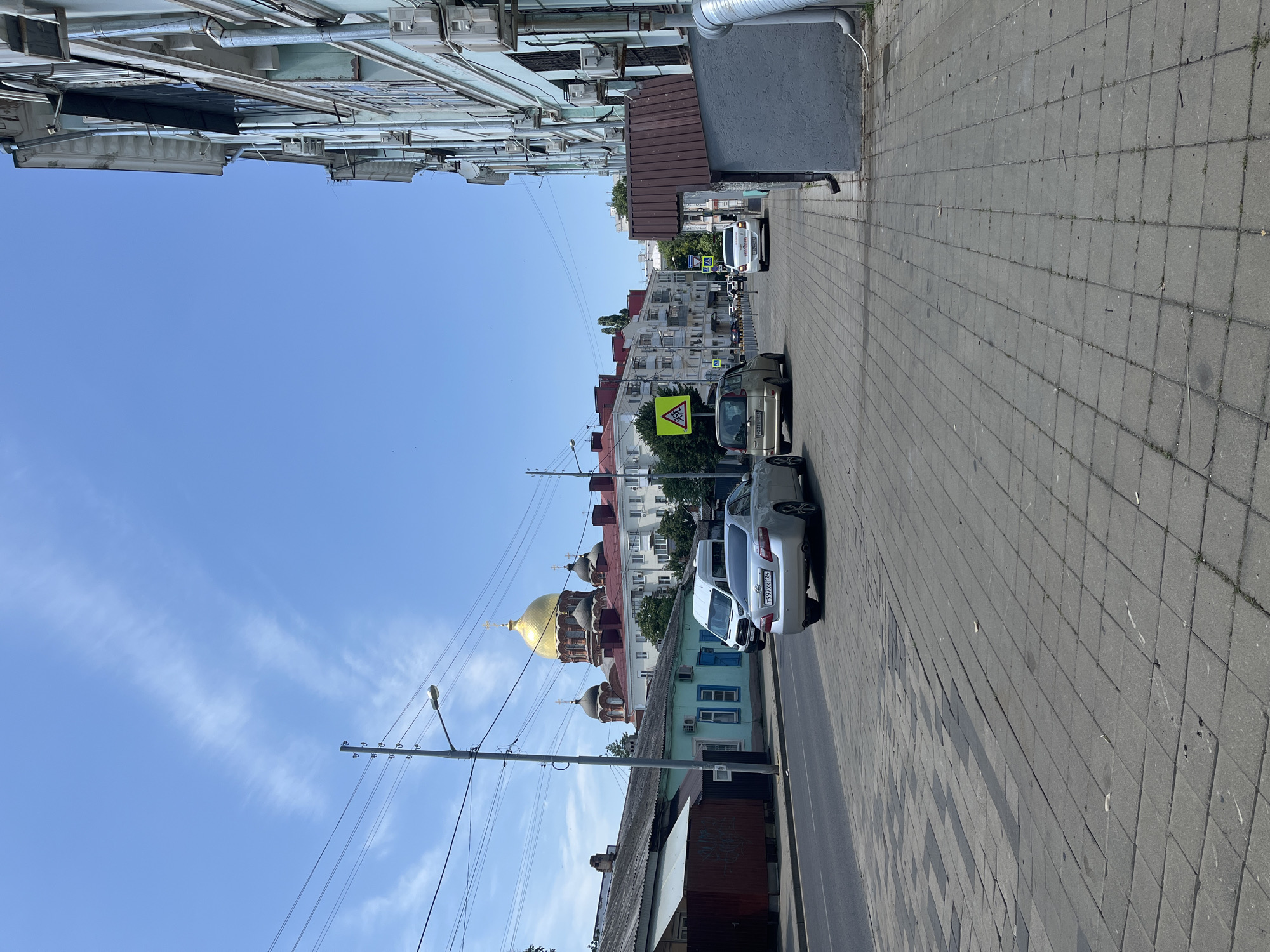 The image depicts a street scene in Krasnodar, Russia, likely on a sunny day with a clear blue sky and some scattered clouds. On the left side of the street, there is a low-rise building with a dark roof and light blue walls, possibly a residential building or a commercial establishment. Further down the street, a large building with a golden dome, characteristic of a church or cathedral, is visible, along with other buildings with red roofs and white facades. In the foreground, the street is paved with gray interlocking concrete blocks, forming a sidewalk. There are several cars on the road and parked along the side, including a silver sedan and a white van. The right side of the street features a multi-story building with a light blue facade, adorned with numerous air conditioning units. The scene suggests an urban environment with a mix of older and newer architectural styles.