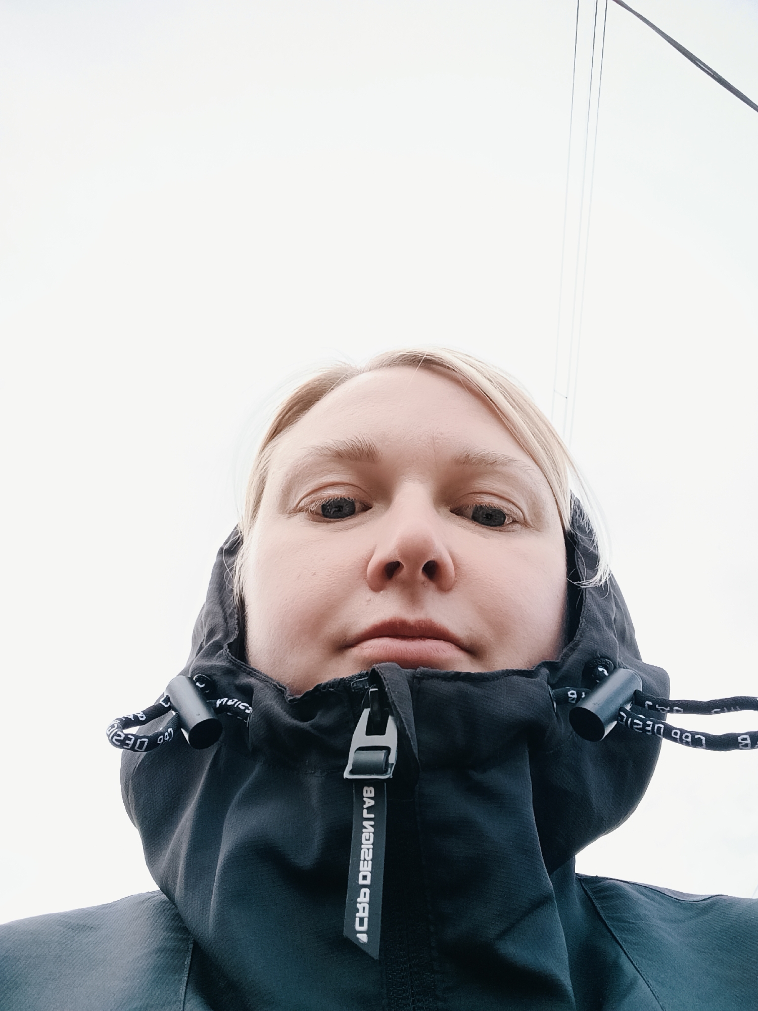 This close-up, low-angle selfie features a person with blonde hair and fair skin, looking directly into the camera with a neutral expression. They are wearing a black hooded jacket, which dominates the lower half of the frame. The jacket has a central zipper, and its hood drawstrings are visible with repeated white text that appears reversed, reading "LBD DESIGN" and "230" or "023". The zipper pull also displays reversed text: "LBD DESIGN RVB". The background consists of a bright, uniformly white sky, suggesting an overcast day. Faint, thin dark lines, possibly wires or rigging, are visible in the upper right portion of the image. The setting is outdoors in Chaikovsky, Russia.
