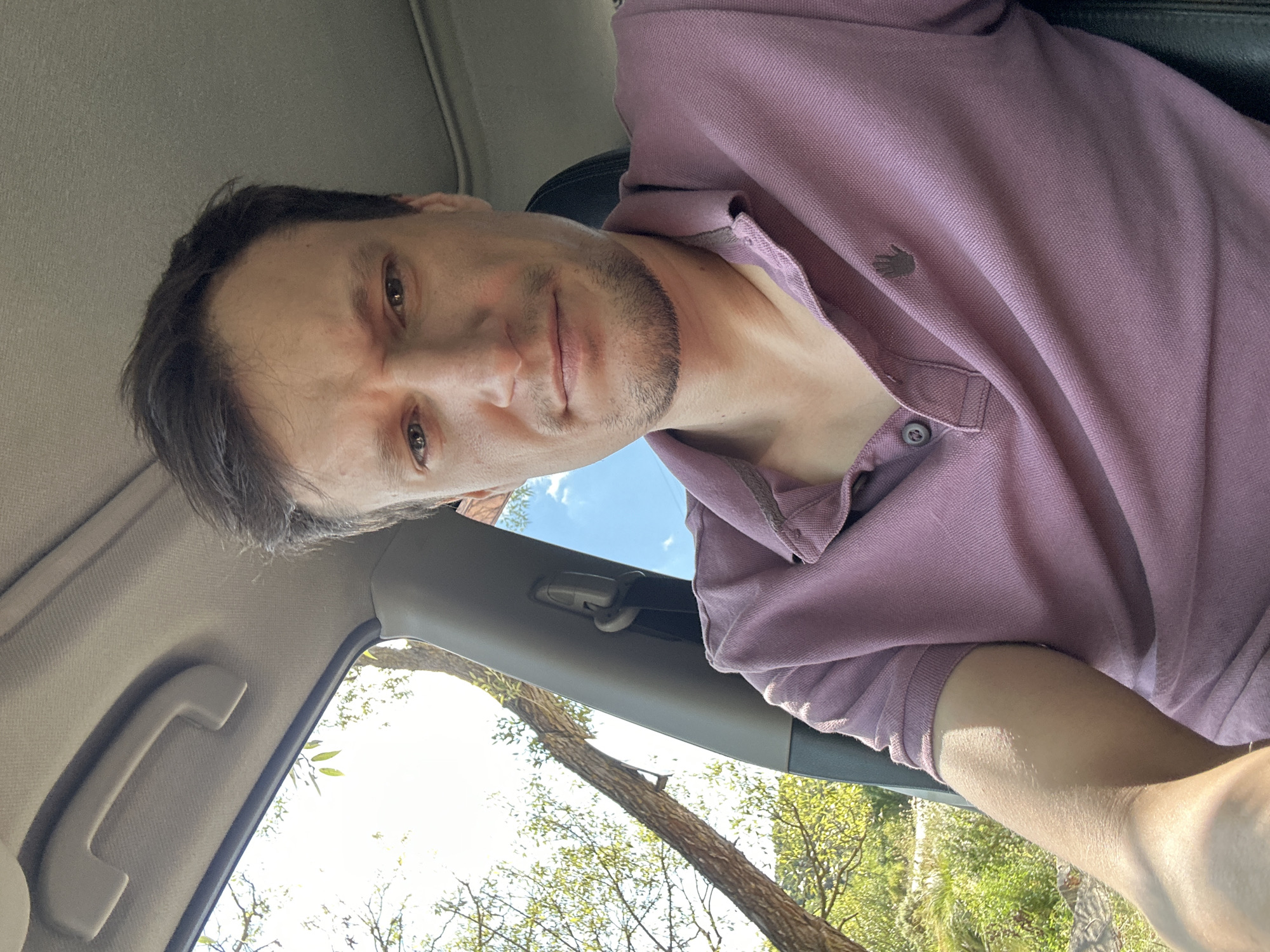 A man is shown from the chest up, inside a vehicle, looking towards the camera. He has short, dark brown hair, a light complexion, and is wearing a light purple polo shirt. The man is seated and the interior of the car is visible, including a grab handle on the ceiling and a seatbelt. Outside the car window, trees and greenery are visible, suggesting an outdoor setting. The image appears to be a selfie taken during the daytime.