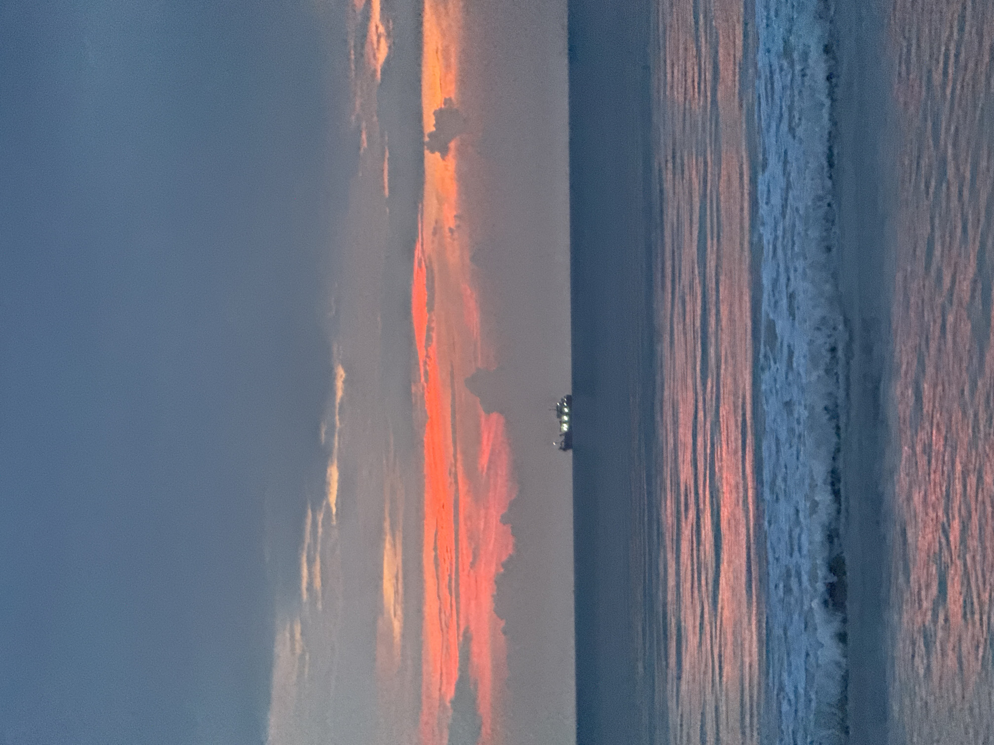 The image depicts a seascape at dusk or dawn. To the left, the sky is a deep blue, transitioning into a gradient of orange and pink hues towards the center. These colors are reflected on the water's surface, creating a shimmering effect. Waves are visible on the right side, marked by foamy white crests. A small, dark object, possibly a boat or ship, is silhouetted on the horizon. Given the location, the scene likely depicts a sunrise or sunset over the Indian Ocean off the coast of Kuta Selatan, Indonesia.