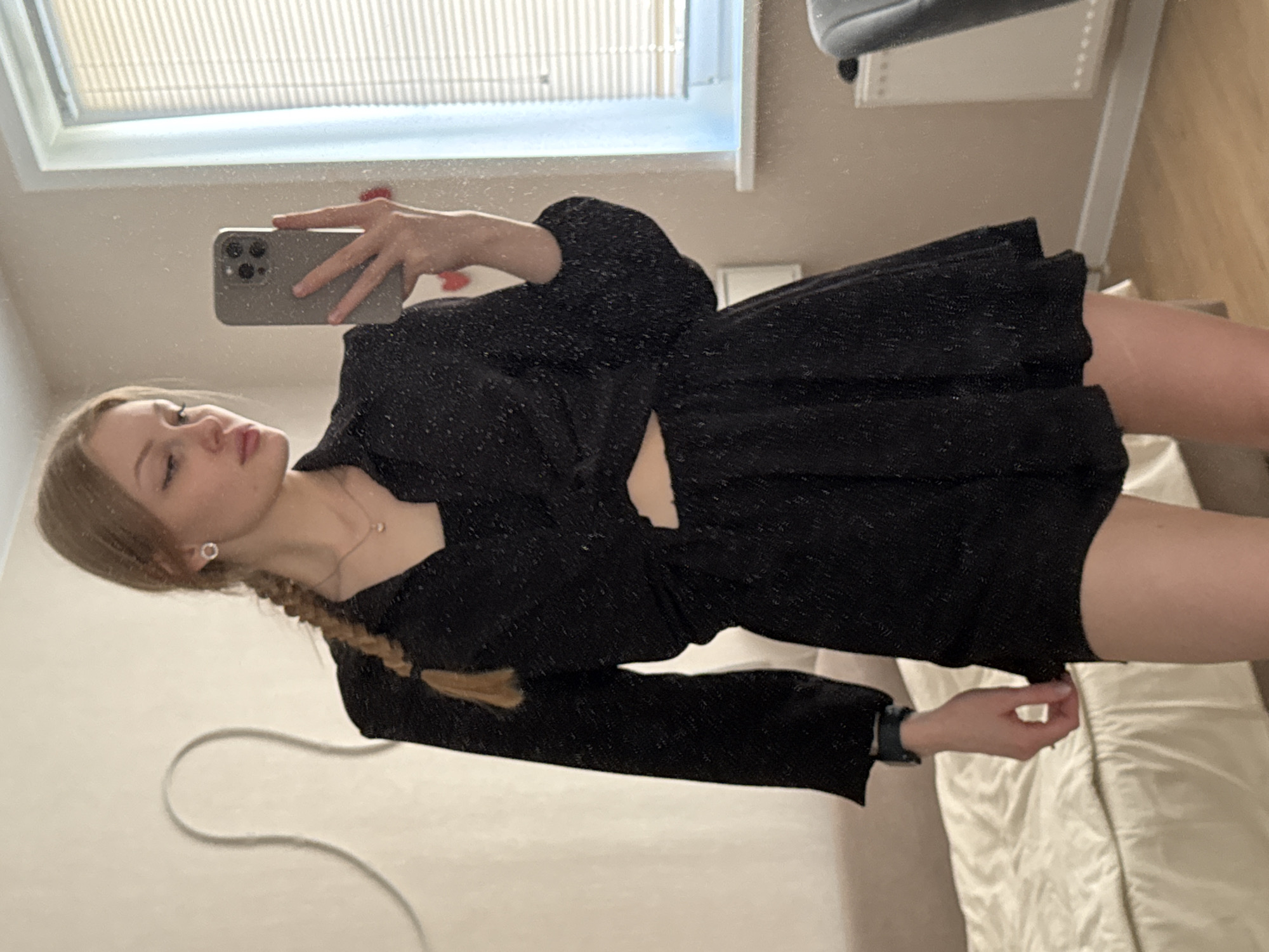 A woman is taking a selfie in a mirror. She has blonde hair styled in a braid and is wearing a black, textured dress with long sleeves. She is holding a smartphone in her right hand. The background includes a window with closed blinds, a piece of furniture (possibly a wardrobe), and a bed with a white comforter. The location is likely indoors, but the specific location in Perm, Russia, cannot be determined from the image.