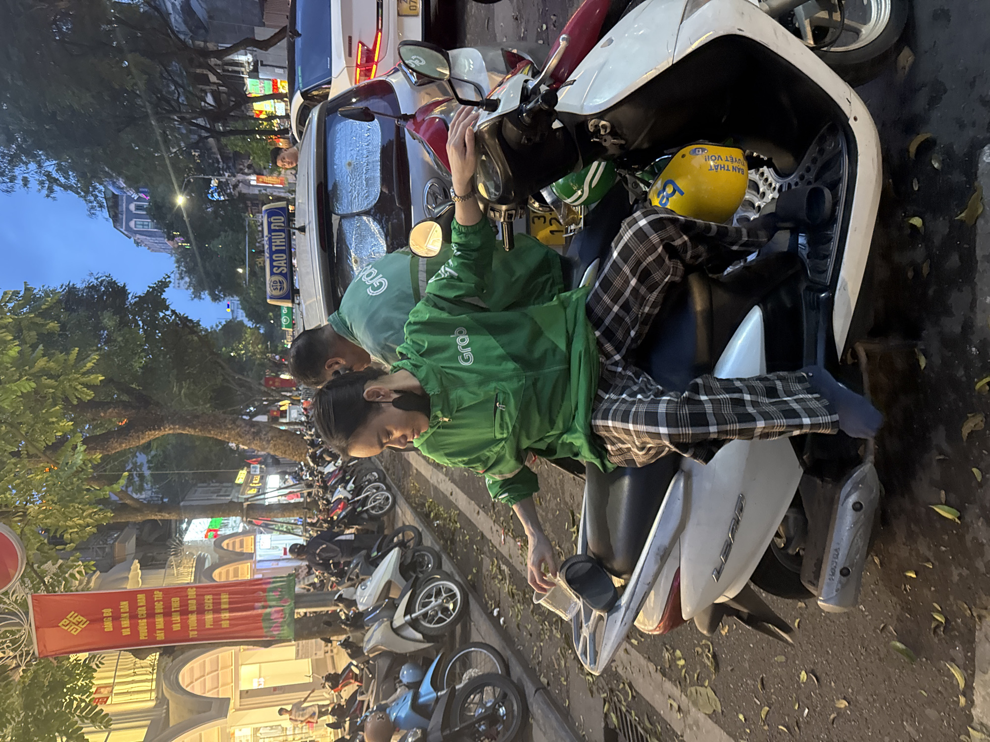 A nighttime street scene in Hà Nội, Vietnam, shows a man wearing a green jacket with "Grab" printed on it sitting on a white Honda Lead scooter. He is wearing plaid pants and appears to be holding something in his hands, possibly related to the scooter. Another man, also wearing a green "Grab" jacket, is standing behind him. Several other parked scooters line the street. A yellow helmet with Vietnamese text is in the scooter's front compartment.
Behind the scooters, storefronts with lights and signs are visible. A red banner with Vietnamese text hangs from a building. A "SAO THỦ ĐÔ" (Capital Star) sign, likely referring to a taxi company, is affixed to a white vehicle parked behind the scooter. The street surface is dark and littered with leaves. Overall, the scene suggests a bustling street corner in Hà Nội with various transportation options.