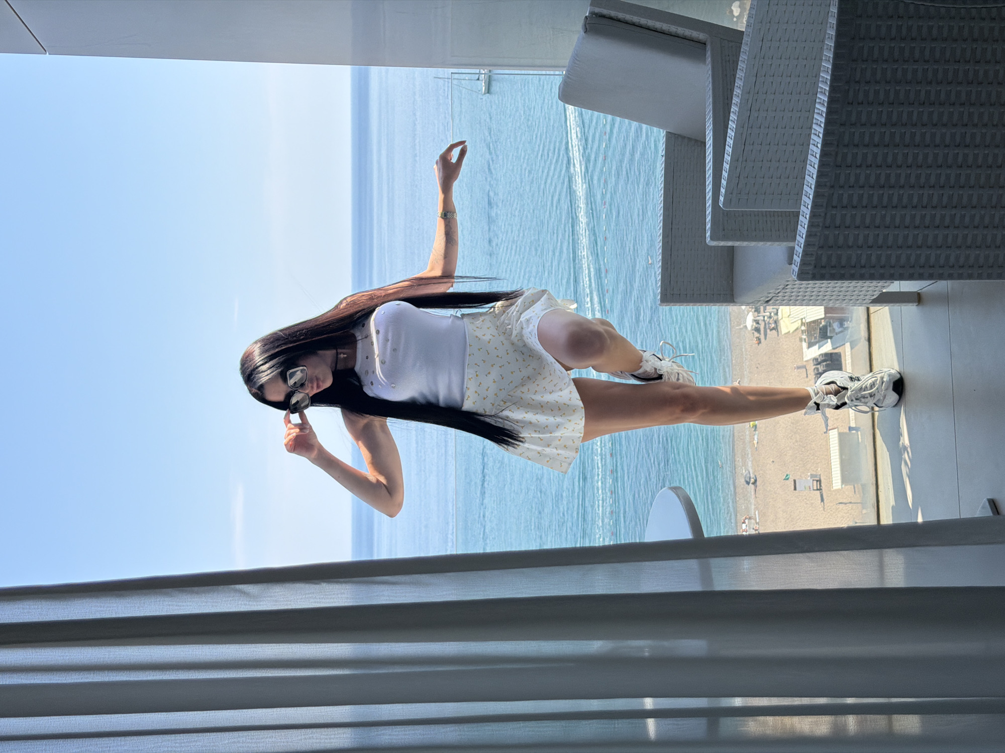 The image depicts a woman with long, dark hair wearing a white top, a patterned skirt, and sneakers. She is also wearing sunglasses and has one arm raised in the air. She is standing on a balcony overlooking the sea and a beach. The location appears to be in Boreti, Montenegro. Wicker chairs are visible on the balcony, with a blue sky and sea in the background, as well as a sandy beach below.