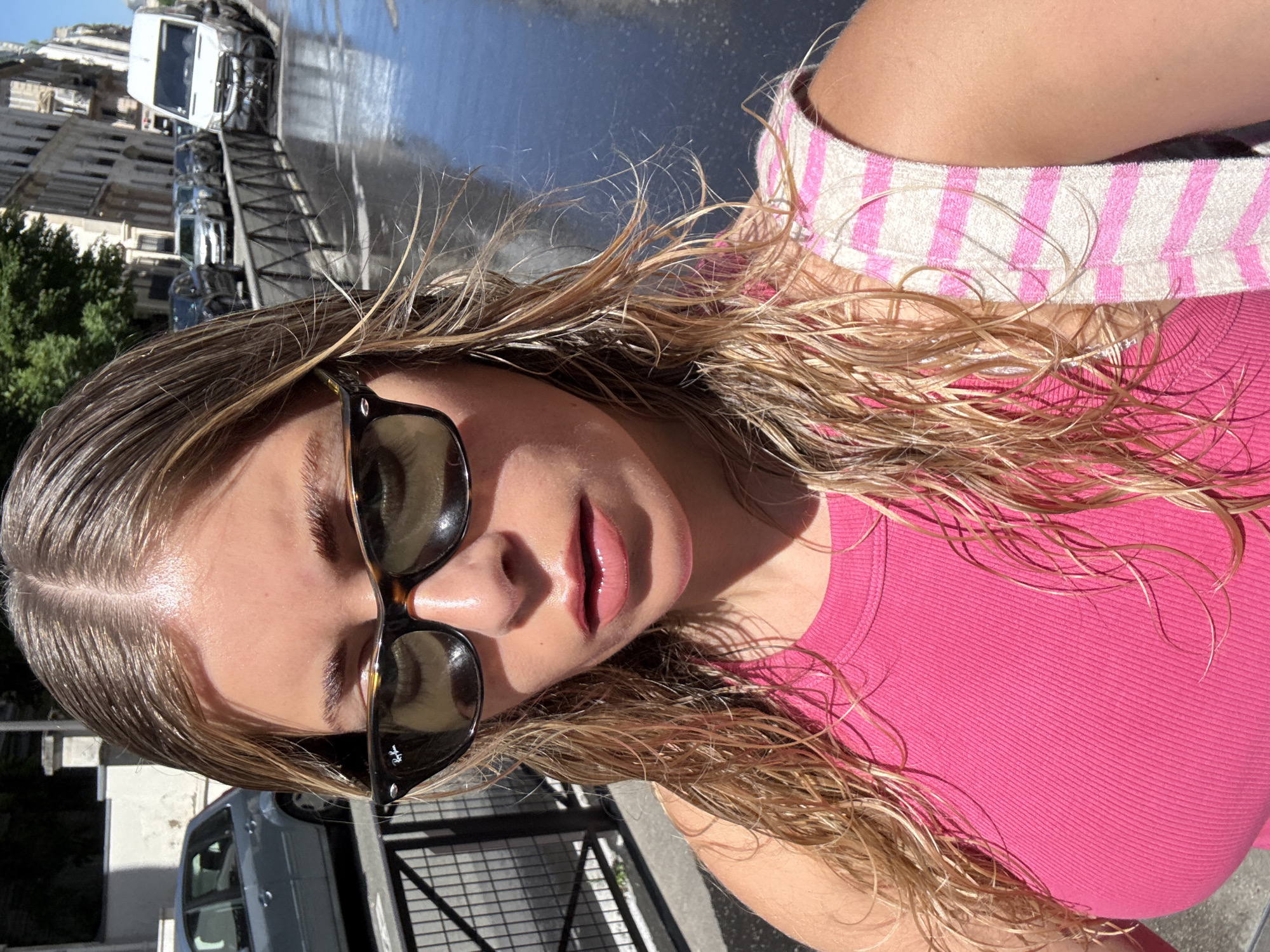 The image is a close-up selfie of a woman with wet, blonde hair. She is wearing sunglasses and a bright pink top with a pink and white striped towel around her shoulders. The background reveals an urban scene, specifically Paris, France, with parked cars, buildings, and trees visible. The wet pavement suggests it may have recently rained or been cleaned.