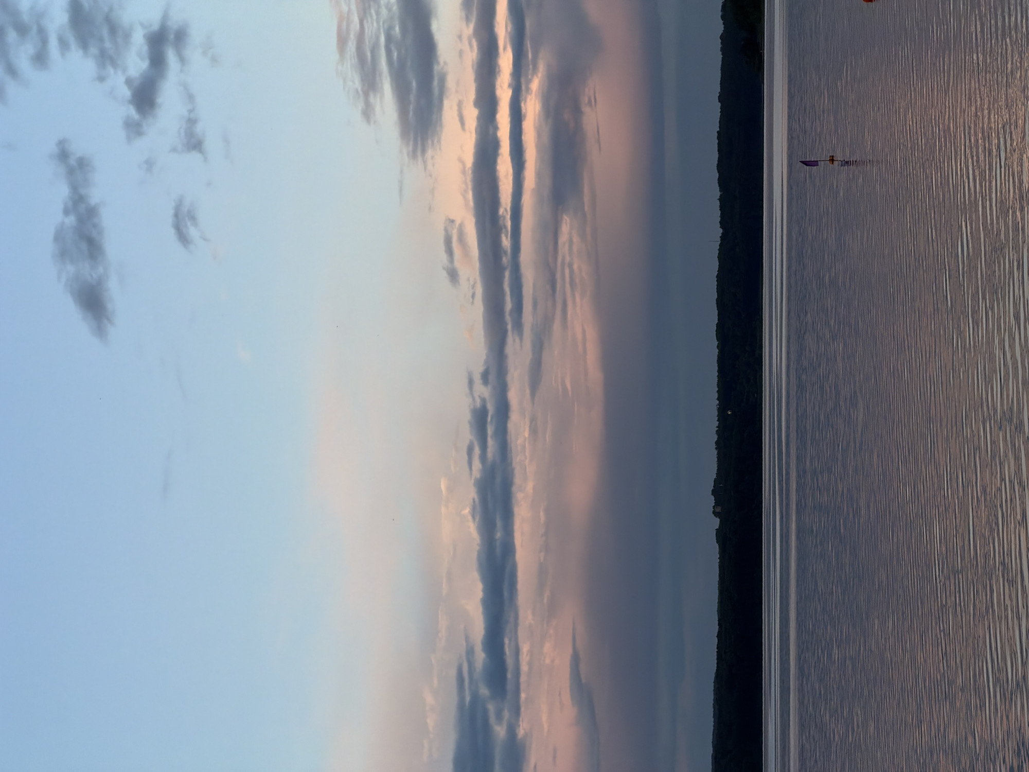 The image shows a serene sunset over a body of water, likely Lake Senezh in Solnechnogorsk, Russia. The sky transitions from a pale blue at the top to hues of pink and purple as it meets the horizon. Wispy clouds are scattered across the sky, some tinged with the warm colors of the setting sun.
The water is calm, reflecting the colors of the sky with gentle ripples disturbing the surface. In the distance, a dark, tree-lined shore is visible, suggesting a natural landscape. A small, purple-sailed object, possibly a model sailboat or buoy, is situated on the water towards the right side of the frame. Further to the right, a small orange object, possibly another buoy, is also visible. The overall impression is one of tranquility and natural beauty at dusk.