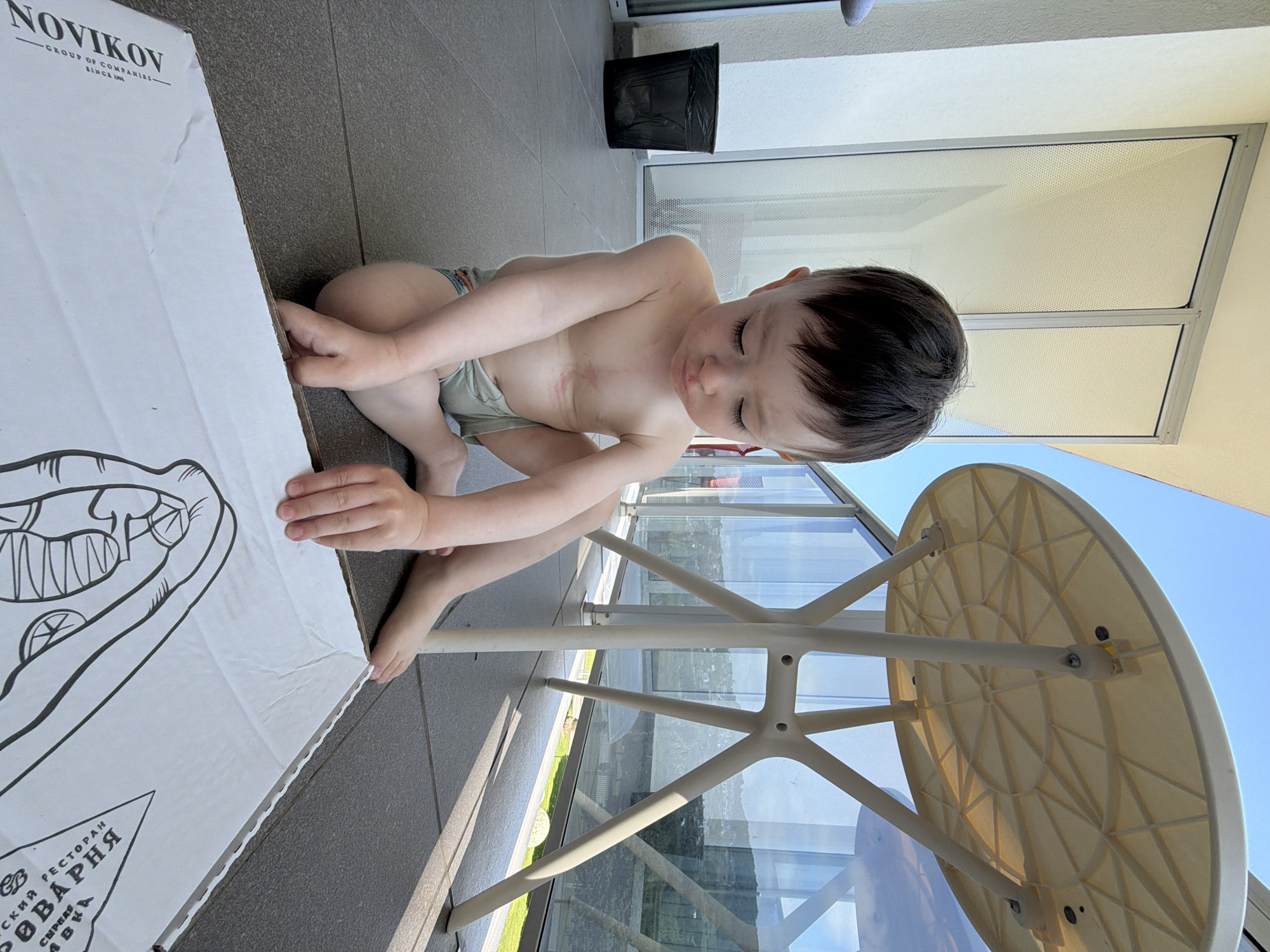 A child sits shirtless on a balcony in Sochi, Russia. The child is drawing on a large sheet of paper with a restaurant's logo and illustration. The balcony has gray tile flooring, glass railings, and a black trash can is visible in the background. A small table with a round top and white legs is in the foreground. The sky is visible behind the glass railing.