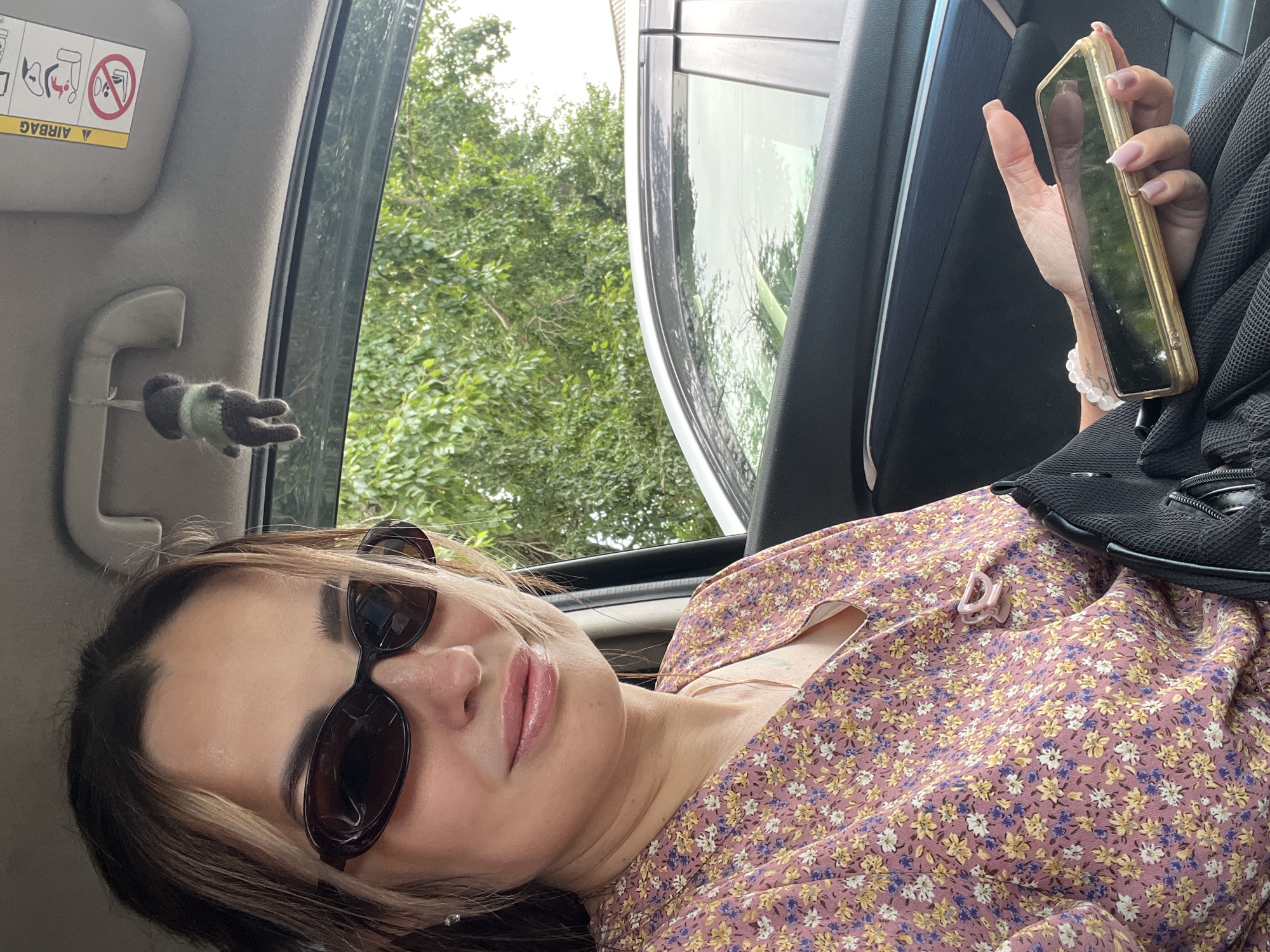 A woman is sitting in the driver's seat of a car. She is wearing sunglasses and a floral-print dress. She is holding a gold-colored mobile phone in her right hand. A knitted doll is hanging from the sun visor above her. The background shows trees and another car. The scene takes place in the city of Unknown, Unknown.