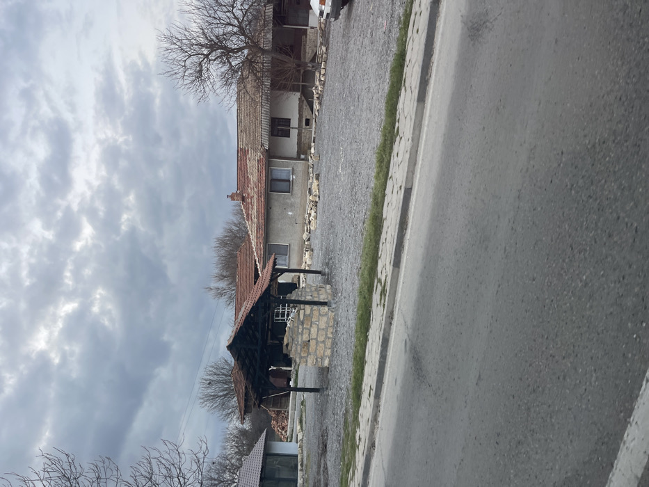 The scene depicts an outdoor environment in Osenets, Bulgaria, under an overcast sky. In the foreground, an asphalt road runs horizontally, bordered on the right by a concrete sidewalk and a thin strip of green vegetation. Beyond the sidewalk lies a large, open plot covered primarily with light grey gravel. Centrally located within this gravel area is a stone well constructed from light-colored, rectangular blocks, topped by a dark wooden canopy with a reddish-brown tiled roof. To the far right, the front bumper and headlight of a white vehicle are partially visible, parked on the gravel. Numerous irregular stones or concrete fragments are scattered across the gravel plot, particularly in front of the main building.
In the mid-background, multiple structures are visible. To the left, a smaller building with a dark roof and large window panels is present. Directly behind the well structure is a house with light-colored walls, multiple windows, and a reddish-brown tiled roof featuring a chimney. To the right of this house, another dwelling appears, distinguished by an open porch or veranda supported by dark pillars and a similar tiled roof. The background horizon features a dense line of bare deciduous trees, indicative of a dormant season. No people or active interactions are observed.