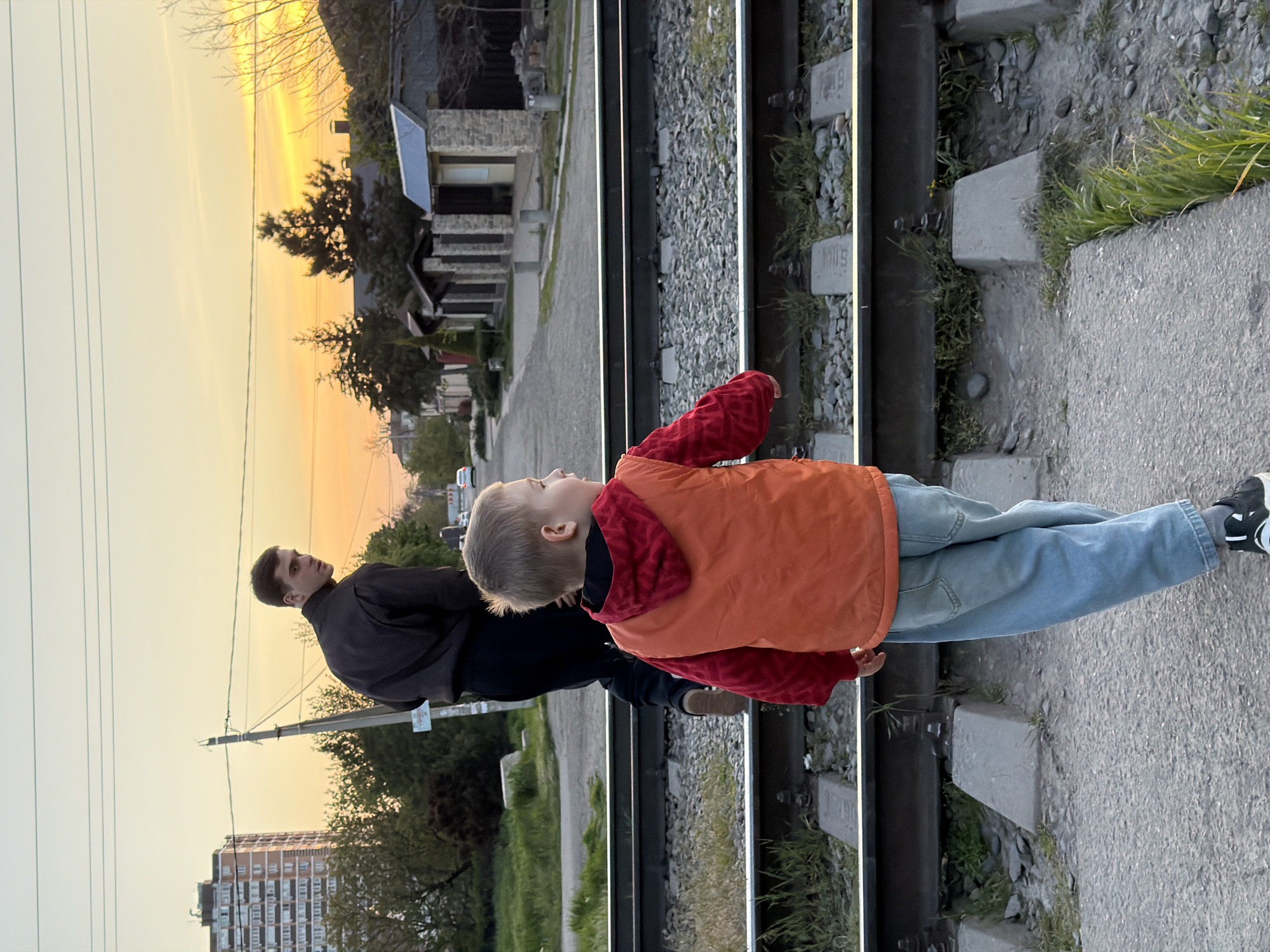 The image captures an adult male and a young boy standing beside railway tracks during what appears to be sunset in Yuzhny, Russia. The man, with short dark hair, is positioned behind and to the left of the boy, wearing a dark top and looking towards the right. The boy, in the foreground, is dressed in an orange sleeveless jacket over a red patterned long-sleeved shirt, light blue jeans, and dark sneakers with white details. He has light-colored hair and is also gazing towards the right, seemingly looking down the railway line.
The setting includes two parallel railway tracks, constructed with gravel and concrete sleepers, running from the foreground into the distance. A paved path or platform borders the tracks on the right side of the frame, where the individuals are standing. Beyond the tracks, a residential area is visible with several houses and a taller, multi-story apartment building in the far left background. Trees and green vegetation are scattered throughout the scene, adding natural elements to the urban-like environment. Overhead power lines crisscross the sky on the left.
The time of day is characterized by a warm, golden sky, indicative of either a sunrise or sunset, casting a soft, orange-yellow glow across the landscape and creating long shadows. This lighting imbues the scene with a tranquil and contemplative mood. On some of the concrete railway sleepers, faded alphanumeric text is visible, with "H118" being clearly distinguishable on at least one block.