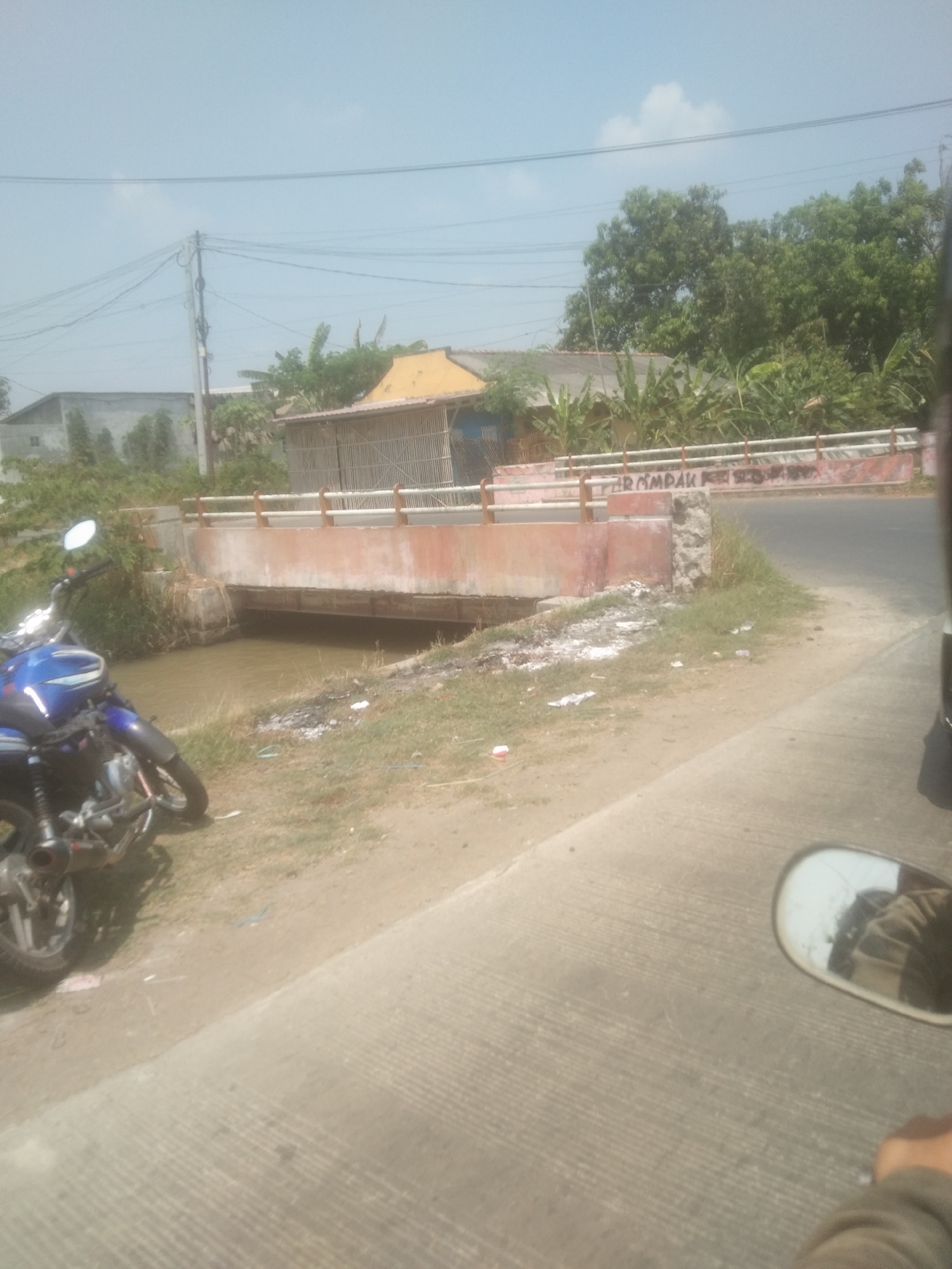 A blue motorcycle is parked on the grassy shoulder of a dirt road next to a river. A bridge spans the river, and in the background, there are houses and trees. The sky is clear and blue. The scene appears to be in a rural area of Desa Cilandak Lor, Indonesia, based on the presence of traditional housing structures and tropical vegetation.