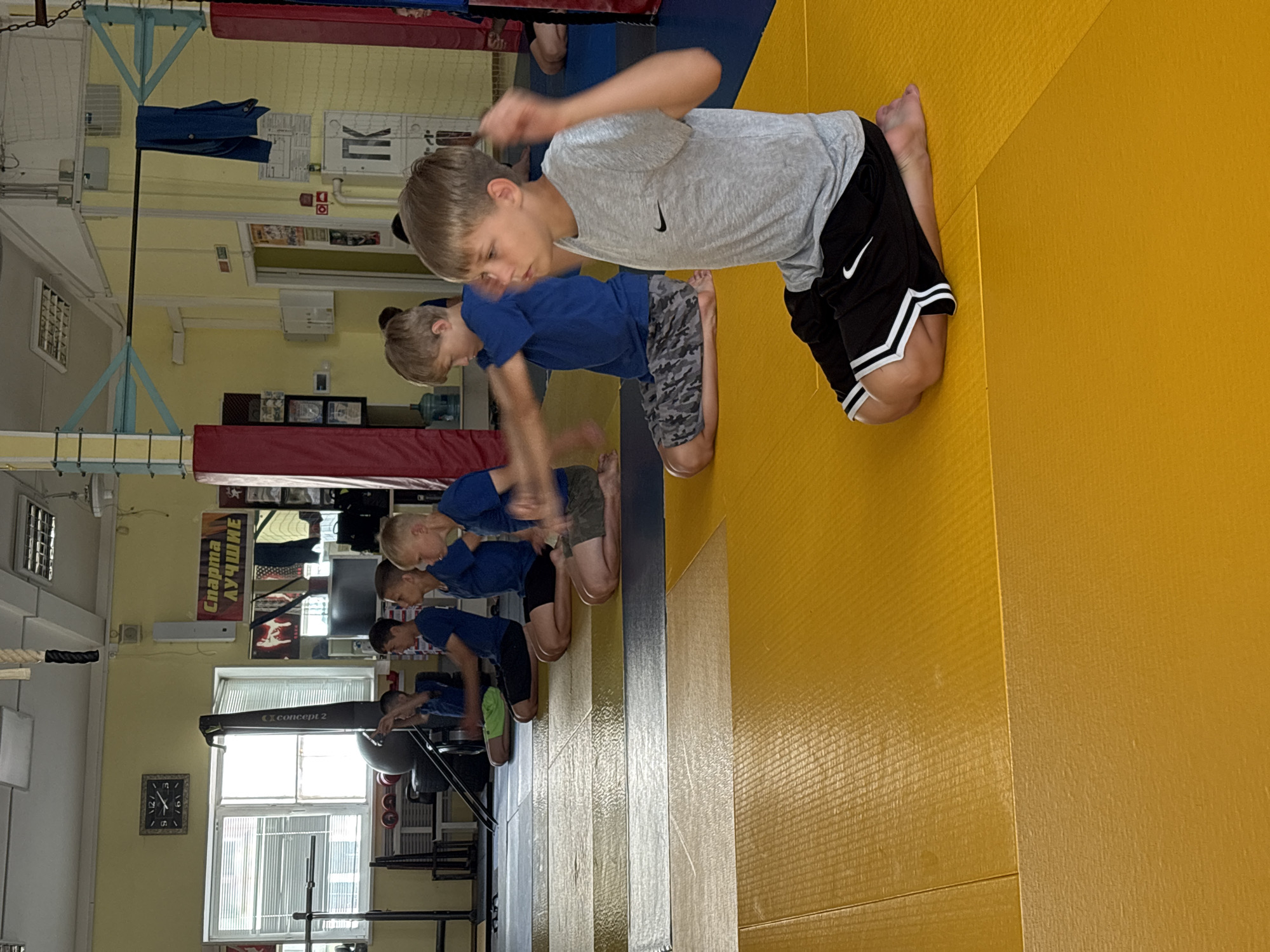 In a gymnasium in Chaikovsky, Russia, a group of young boys are performing exercises on a yellow mat. The boys, dressed in athletic wear, are positioned in a seated or kneeling posture and appear to be engaged in a synchronized movement, possibly a form of martial arts training or warm-up. The gymnasium environment is visible with gym equipment such as a rowing machine, weights, and climbing apparatus in the background. A clock is mounted on the wall, displaying the time as approximately 12:10. Motivational posters, including one with the Russian text "Спарта Лучшие" (Sparta Best), are also present on the walls. The overall scene depicts an active training session within a sports facility.