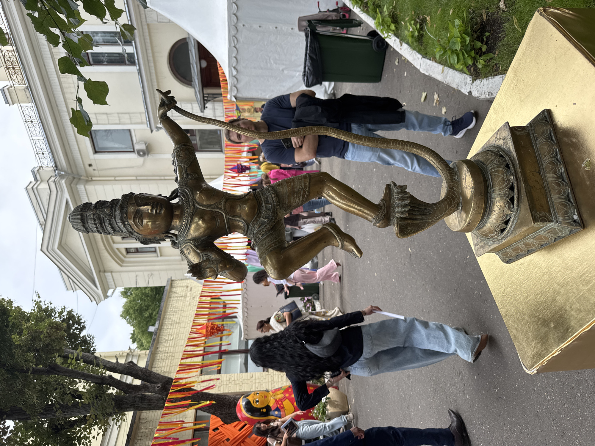 The image depicts an outdoor scene in Moscow, Russia, likely at a cultural festival, given the presence of colorful decorations. In the foreground, a bronze statue of a dancing deity, possibly Shiva, stands on a golden pedestal. The statue is intricately detailed, with the deity in a dynamic pose, holding a serpent. Behind the statue, several people are visible. One woman in the mid-ground with long dark hair wears a black top and light-wash jeans. Another man, standing further back to the right, is wearing a dark blue t-shirt and light blue jeans. The background shows a building with classical architecture, possibly a museum or cultural center, with tents and more decorations suggesting a festive event. The ground is paved, and there is greenery and a trash receptacle on the right side of the image.