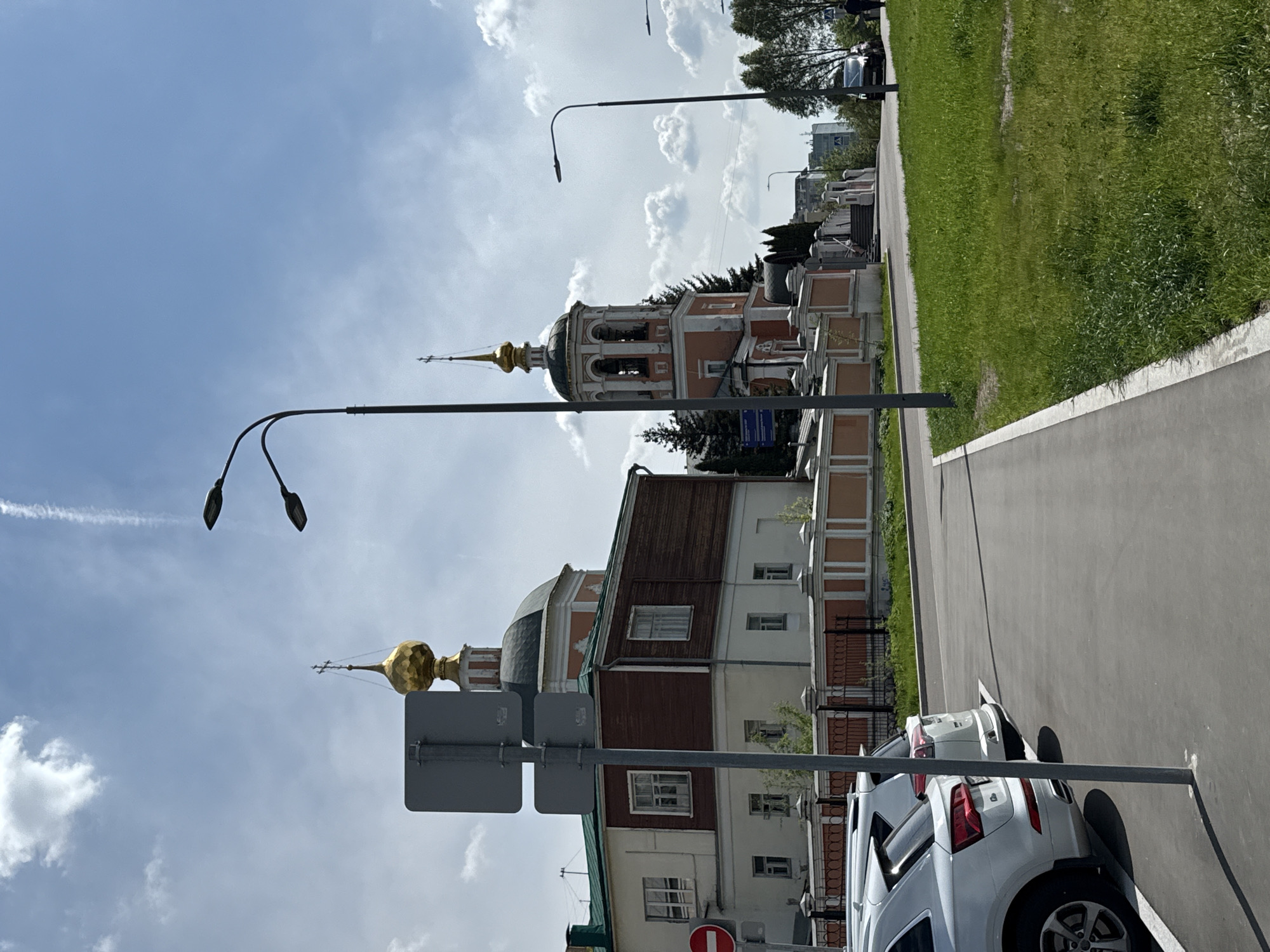 The image depicts a street scene in Moscow, Russia, featuring the Donskoy Monastery. Visible are the monastery's buildings with golden domes and detailed architectural elements. A street runs along the side, with parked cars and lampposts. Across the street, there's a cemetery with headstones and greenery. The sky above is partly cloudy.