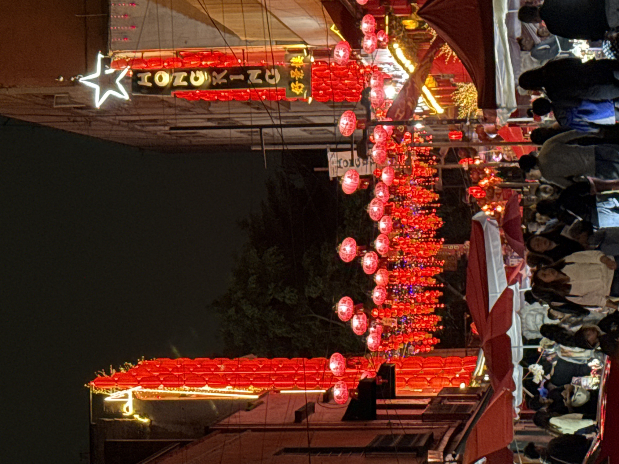 At night, a street in Mexico City is packed with people. Red paper lanterns hang densely above the street, and some people are holding umbrellas. A large sign with Japanese characters and a star symbol is visible. The scene conveys a bustling urban atmosphere, likely during a festival or special event.