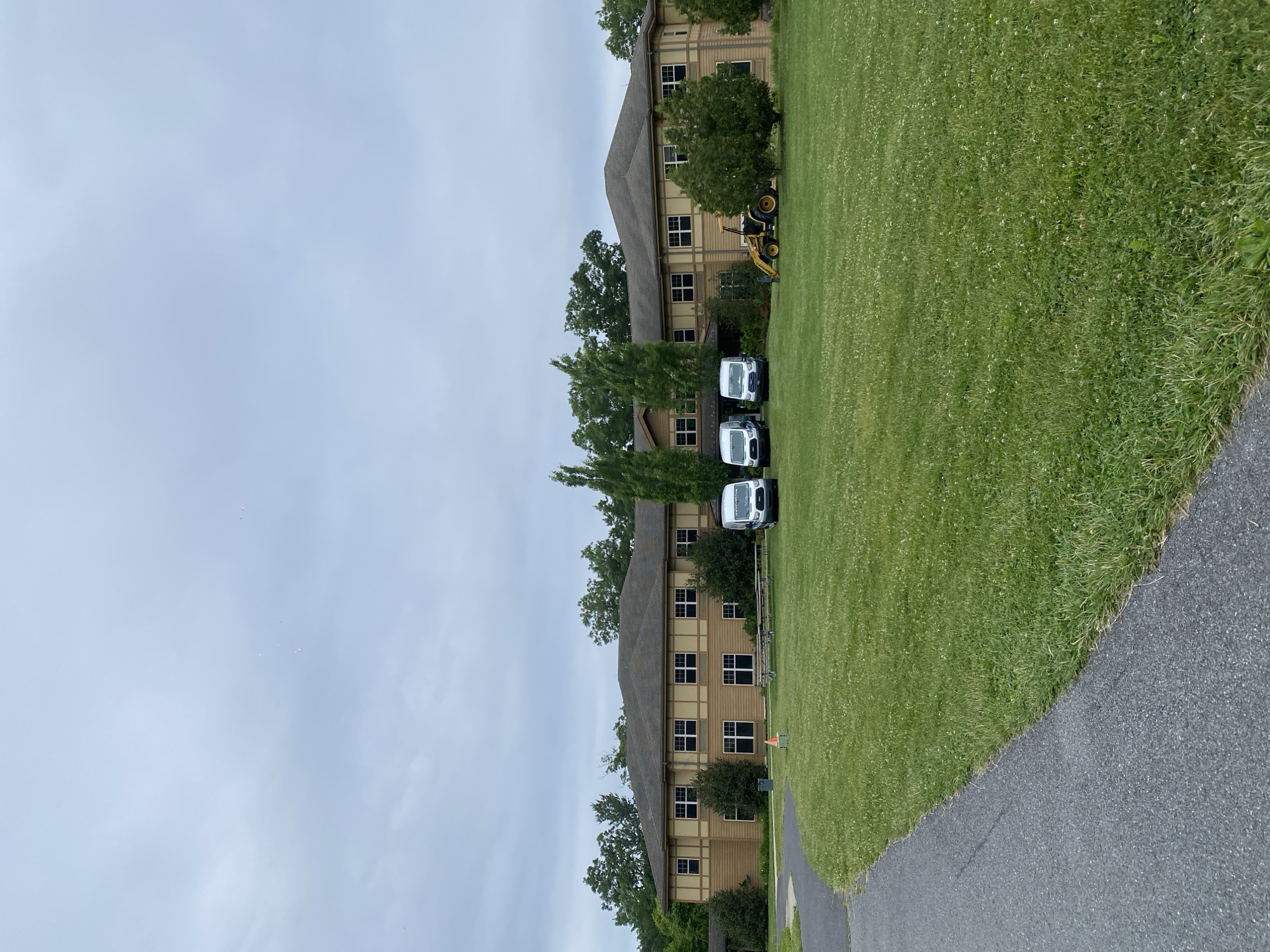 In Washington Township, United States of America, on a cloudy day, a two-story building with beige siding and dark brown trim stands in the background. The building has numerous evenly spaced windows with dark frames. A dark asphalt pathway curves into the foreground from the left. A large, well-maintained lawn with scattered white flowers surrounds the building. Three white panel vans are parked in front of the building, and a yellow backhoe is positioned to the right of the vans. Trees and shrubbery are visible behind and to the sides of the building, with a dense line of trees forming the background. The sky is overcast with faint pinkish specks visible high up.