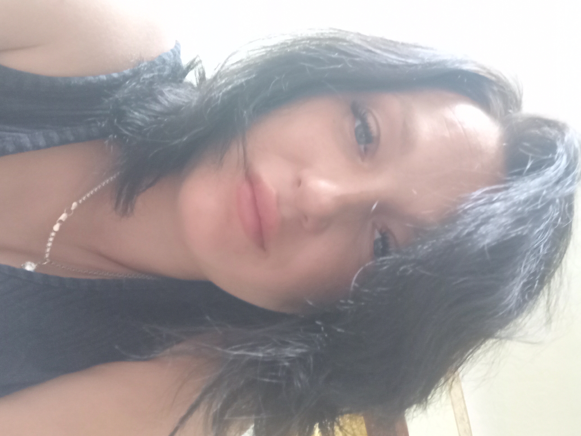 The image is a close-up, selfie-style photograph of a woman with dark, wavy hair. She is wearing a dark blue, sleeveless top and a silver necklace with a pendant. Her eyes are open, and she appears to be looking directly at the camera. Her lips are slightly parted. The lighting in the image is bright, with a light source seemingly coming from the left side, creating some blown-out highlights. The background is mostly out of focus and appears to be a wall or interior space, with some indistinct elements visible on the right side, possibly a wooden surface. There are no discernible elements in the image that would allow for the determination of its location in Chuhuiv, Ukraine, or any urban environment.