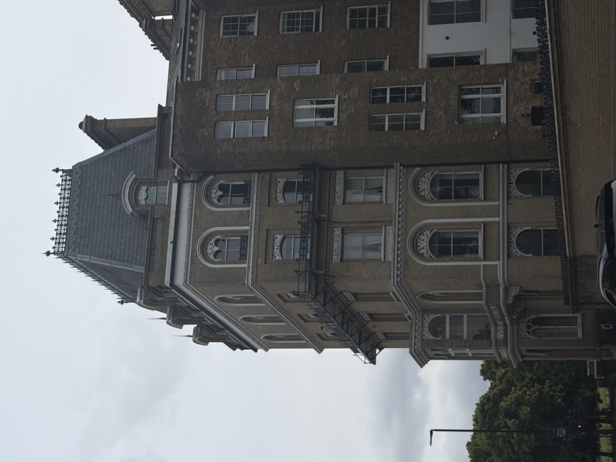 The image captures a multi-story, ornate building under an overcast sky, located in South Kensington, London, United Kingdom. The prominent structure on the left features a light tan brick or stone facade and is topped by a steeply pitched dark grey mansard roof, adorned with decorative black iron cresting along its ridge and several arched dormer windows. The building's floors are punctuated by numerous arched windows with intricate white decorative surrounds, some equipped with small, dark-colored wrought-iron balconies.
To the right, an adjacent building, constructed of darker brown brick, presents a plainer architectural style with multiple rectangular sash windows of varying dimensions. Visible in the foreground is a low brick wall, behind which the top portion of a dark-colored vehicle is partially discernible. On the far left, a black street lamp stands next to a circular red and white speed limit sign indicating "20." Behind these, a dense canopy of green trees suggests a park or green space bordering the road. No people or specific actions are visible in the scene.