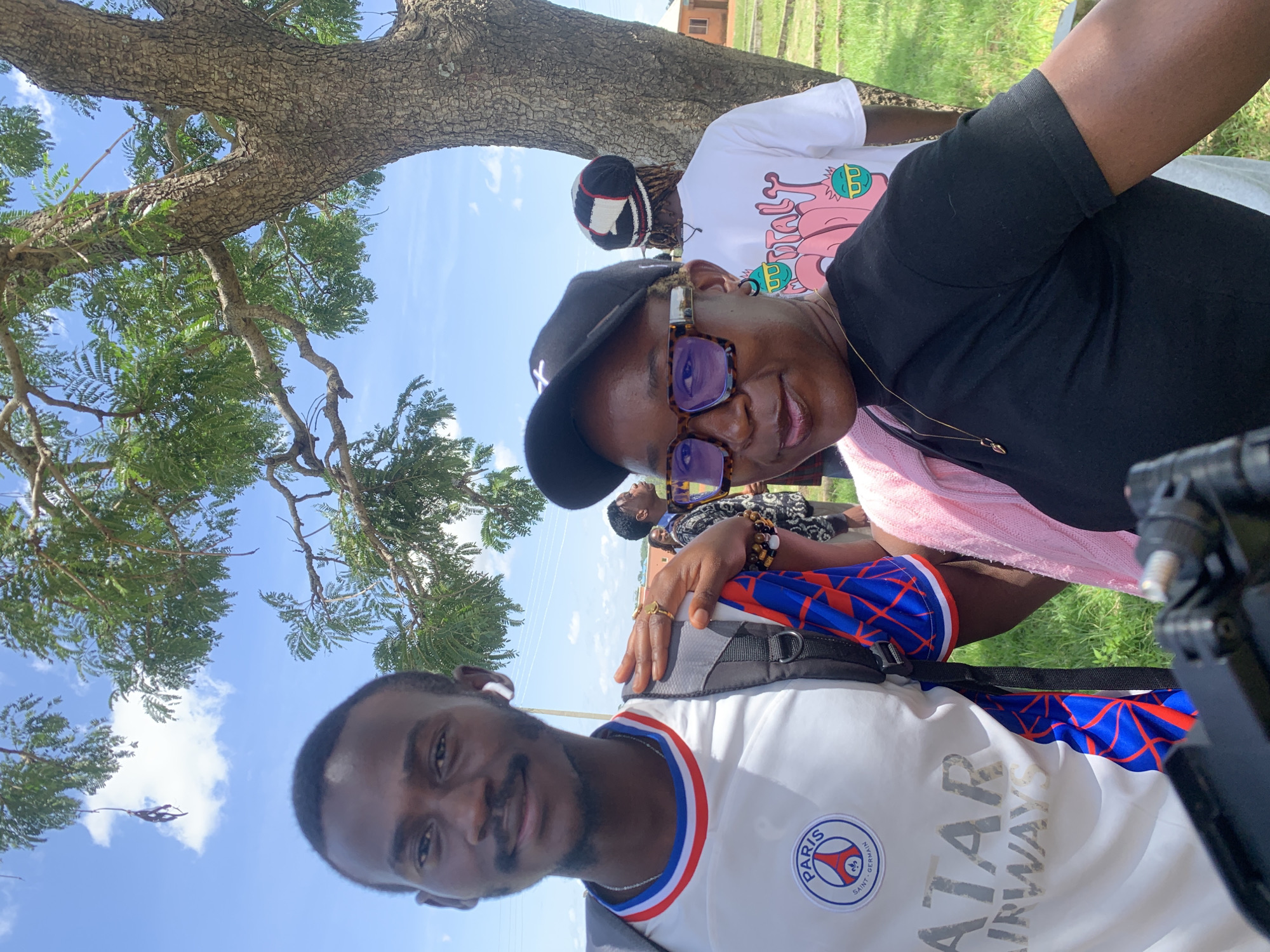 In Gwafan, Nigeria, a young Black man and woman are posing for a selfie outdoors. The man, positioned slightly to the left, wears a white Paris Saint-Germain jersey with a blue and red accent on the collar and sleeves, and a backpack. The woman, to the right, sports a black t-shirt, a black baseball cap, and tortoiseshell glasses. She has a pink cloth draped over her shoulder. In the background, another person wearing a white t-shirt with a colorful graphic and a beanie is visible, along with hints of buildings and greenery. The scene is set under a tree with green leaves, against a bright blue sky with scattered clouds.