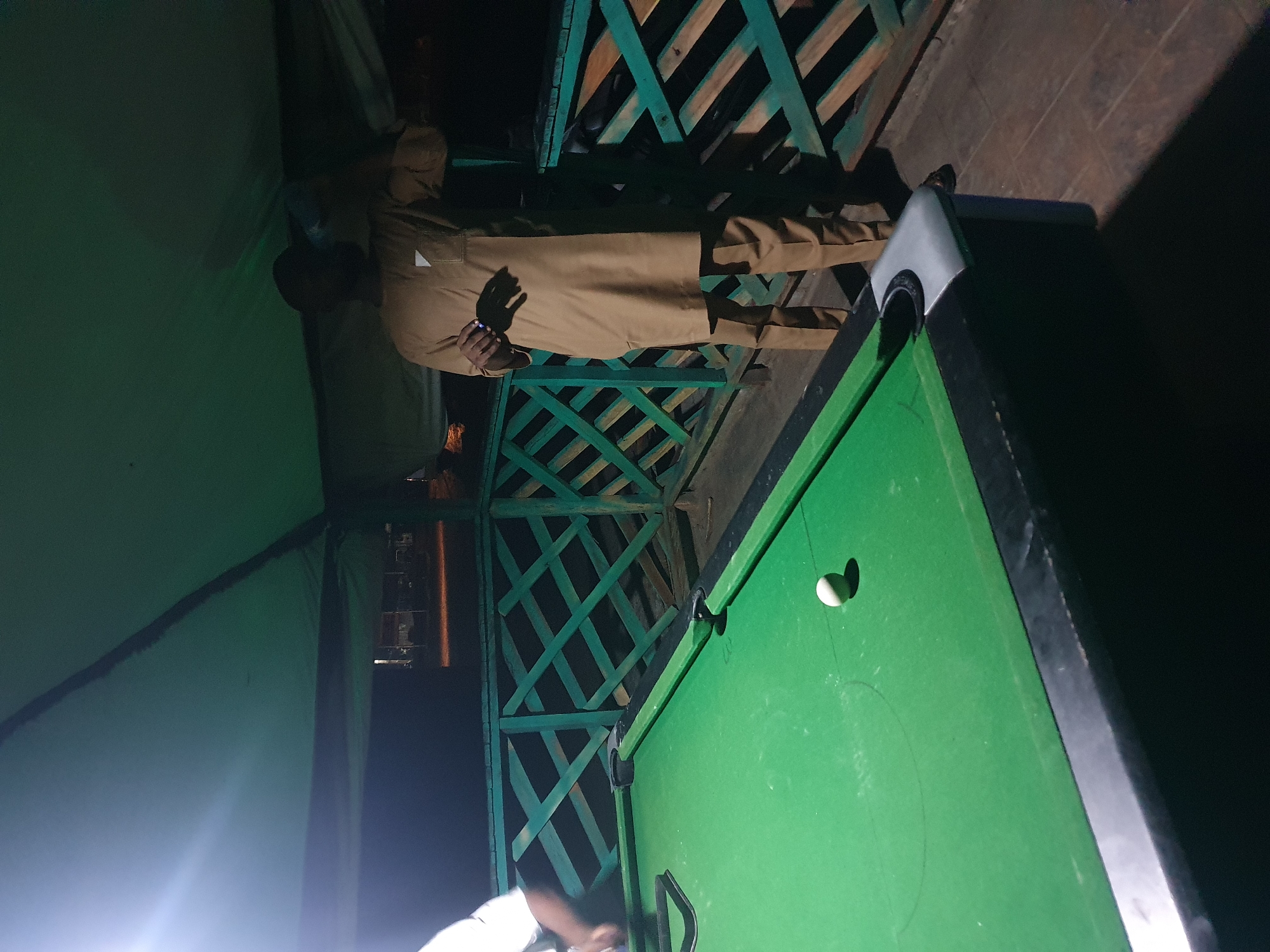 The image depicts an outdoor area, likely a patio or balcony, in Lokoja, Nigeria. A green billiard table occupies the foreground, with a single white billiard ball positioned on its surface. A man, wearing a light brown traditional outfit, stands to the right of the table, holding a blue object in his hand and appearing to drink. Behind the table, a green wooden railing with a diamond pattern encloses the area, and beyond that, the darkness suggests it is nighttime. The overall environment is dimly lit, with artificial lighting casting shadows and highlighting certain areas.