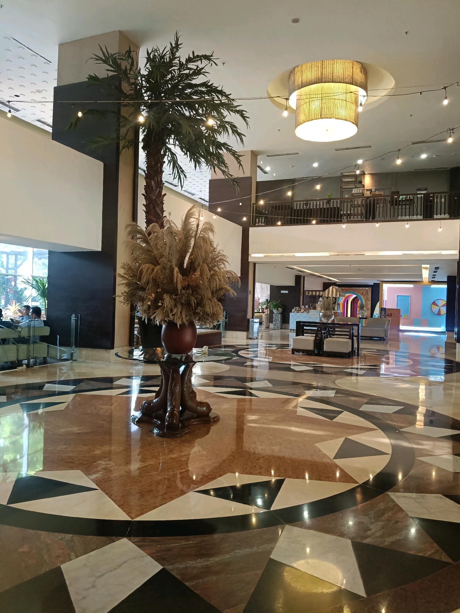 The image depicts the spacious and brightly lit interior of a hotel lobby in North Jakarta, Indonesia. A large, decorative palm tree stands prominently in the center, reaching towards the high ceiling. In front of the palm tree, a vase filled with dried reeds rests on a table with a glass top and a unique pedestal featuring sculpted fish. The floor is a striking mosaic of marble tiles in various shades of brown, beige, and black, arranged in a geometric pattern.
In the background, a multi-story structure with balconies and railing is visible, suggesting a larger building. A large, cylindrical pendant light fixture hangs from the ceiling, casting a warm glow. String lights are also visible, adding to the ambient lighting. To the left, several people are seated in a lounge area near a large window, some appearing to be engaged in conversation. Further in the background, a colorful, abstract mural or installation is visible, adding a touch of vibrancy to the scene. The lobby appears clean and well-maintained, with a sense of modern elegance.
