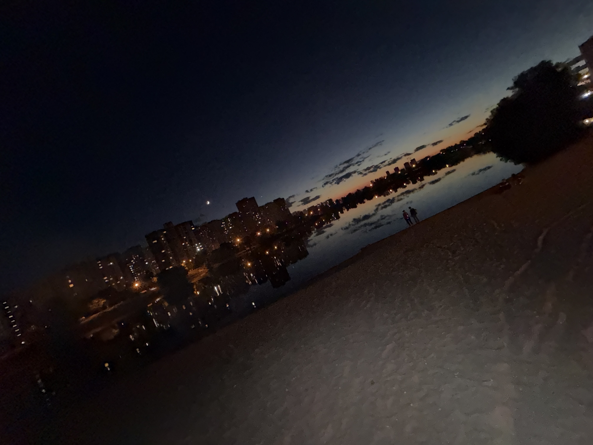 The image depicts a waterfront scene at dusk in Kyiv, Ukraine. The sky is a deep blue, fading to orange and pink near the horizon where a few scattered clouds are visible. A crescent moon is present in the upper left portion of the sky. In the middle ground, a city skyline with illuminated apartment buildings is reflected in the calm water. The foreground consists of a sandy beach. Two individuals, silhouetted against the twilight, are standing on the beach, facing the water. The overall impression is of a peaceful evening by the water in an urban setting.