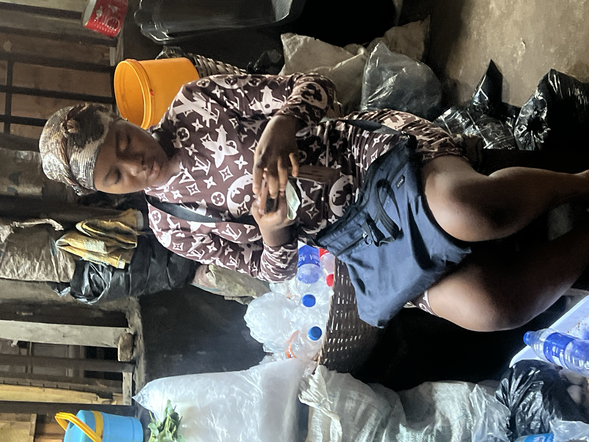 An adult female is shown seated within a cluttered environment in Nkpor, Nigeria. The woman is wearing a brown and white patterned shirt and a headscarf. She appears to be counting money. The woman is surrounded by refuse, including water bottles in a basket, plastic bags, an orange bucket, and several black trash bags. The setting suggests a marketplace or informal business area.