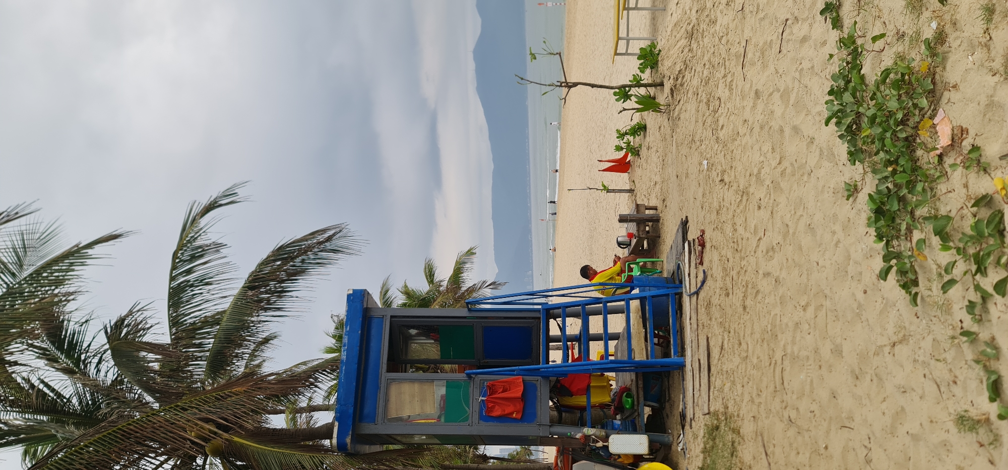 This photograph captures a scene at a beach in Đà Nẵng, Vietnam. The viewpoint is from a low angle, looking up and along the beach. A blue and gray lifeguard tower sits prominently in the foreground, framed by palm trees. A male lifeguard wearing a red and yellow uniform is seated on a green chair next to the lifeguard tower, seemingly at rest or on break. Further along the beach, partially obscured by the tower, are red flags planted in the sand and several small trees, indicating the swimming area. The beach itself is a sandy expanse leading to the sea, where a few people can be seen in the water. Distant mountains are visible against a cloudy sky, setting the backdrop for the beach. Sand and vegetation line the right edge of the photo.