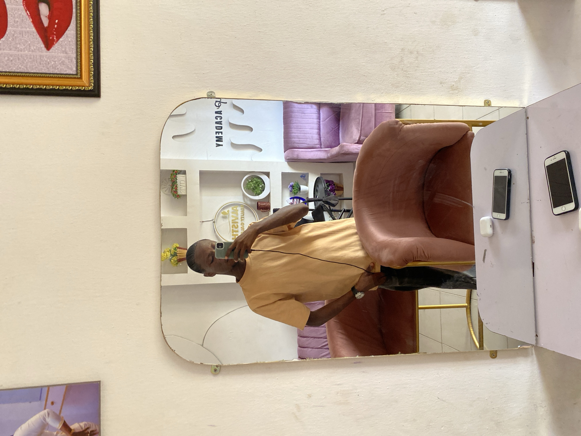 The media file captures a man taking a mirror selfie inside what appears to be a salon or beauty academy in Bukuru, Nigeria. The image is oriented vertically (rotated 90 degrees clockwise).
The main subject is a man, reflected in a large, irregularly shaped mirror, holding a smartphone to capture his image. He is wearing an orange short-sleeved t-shirt, dark trousers, and a watch on his left wrist. His face is partially obscured by the phone.
The mirrored reflection reveals the interior setting:
- To the left, a purple, tufted upholstered sofa or chair is visible.
- Below the sofa, a set of white shelves holds various decorative items, including a potted green plant, another pot with purple flowers, and a round, possibly metallic, object.
- To the right of the man's reflection, a brown upholstered chair is prominent.
- On a white surface next to the brown chair, two smartphones and a white AirPods case are resting.
- The floor visible in the reflection is tiled, and there's a gold-colored metal frame, likely part of furniture.
Visible text in the mirror, appearing reversed and upside down, includes "YMEDACA" (likely "ACADEMY") and "TIWIAZ YTUAEB" (likely "BEAUTY ZAWIT"), which confirms the nature of the establishment. Another small decorative item on a shelf reads "HOME".
Outside the mirror's reflection, on the light-colored wall, a framed picture featuring glossy red lips on a patterned background, set in an ornate gold frame, is visible. Part of another framed image is seen below it. The overall setting suggests an indoor commercial space, likely a beauty or hair salon, during daytime hours, though no specific time or weather cues are present. The man's expression is not discernible due to the angle and phone.