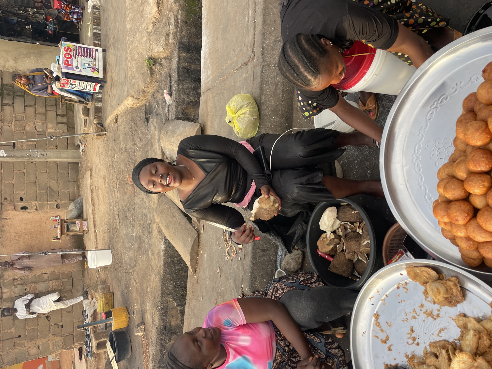 The image depicts a street scene in Jos, Nigeria, with three women engaged in activities related to food vending. In the foreground, two women are seated. One woman on the left, wearing a pink and blue tie-dye t-shirt and a patterned skirt, is looking towards the right. The woman to her right, wearing a black dress and a headscarf, is actively peeling a piece of yam with a knife. In front of them are two large metal trays; one contains fried snacks, and the other holds a pile of small, golden-brown fried dough balls. A black basin filled with chunks of yam is also visible.
Behind the seated women, a third woman, also in black attire, is crouched down and reaching into a white bucket with a red lid. She has her hair braided. Further in the background, two men are standing near a brick wall. One man on the left wears a white traditional outfit, while the other wears a brown garment. A small table with various items, including bottles and what appears to be a POS machine with promotional signage, is set up against the wall. The environment appears to be an outdoor market or roadside stall, characterized by dirt and concrete ground, brick structures, and various containers and equipment typically found in such settings. The lighting suggests it is daytime, possibly late afternoon, given the shadows.