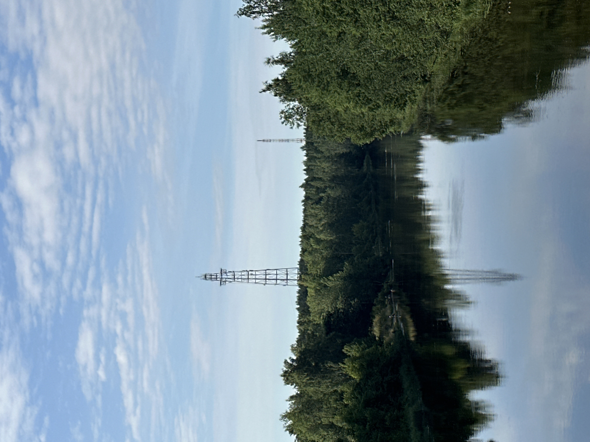 The image depicts a calm, wide river flowing through a lush green forest. Two tall, metallic radio towers are visible on opposite banks, rising above the dense tree line. The sky is a clear blue, dotted with scattered white clouds. The river's surface acts as a mirror, reflecting the trees, towers, and sky, creating a symmetrical visual. On the left bank, a small group of people are gathered near the water's edge, appearing to be enjoying the natural scenery. The overall impression is one of a tranquil, rural landscape with industrial elements integrated into the environment. The location is likely the Luga River, near the city of Slantsy, Russia, given the presence of radio towers and the characteristic riverine landscape.