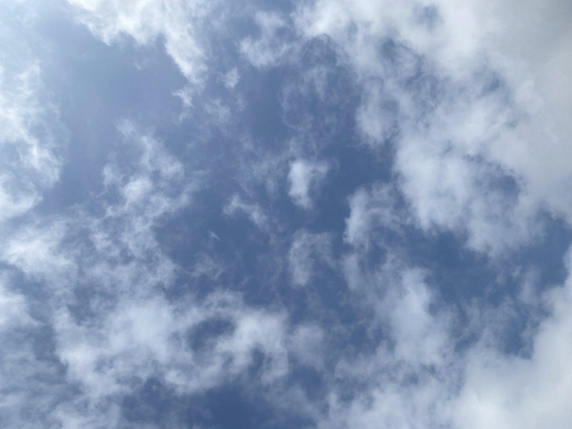 The image is a close-up, upward-facing shot of a partly cloudy sky. The dominant colors are blue from the sky and white from the clouds. The clouds are scattered and appear to be of the cumulus type, with varying densities and shapes. There is a slight haze or lens flare effect visible, which softens the details of the clouds and sky. The overall impression is of a bright, sunny day with some cloud cover.
Based on the provided information, this image depicts the sky above Lafia, Nigeria. The scene is devoid of any specific landmarks or identifiable features that would pinpoint a precise location within the city. It is a generic depiction of the sky as seen from ground level within Lafia. There are no people, subjects, objects, actions, or interactions visible in the image, only the natural phenomenon of clouds against a blue sky. The environment is limited to the sky itself.