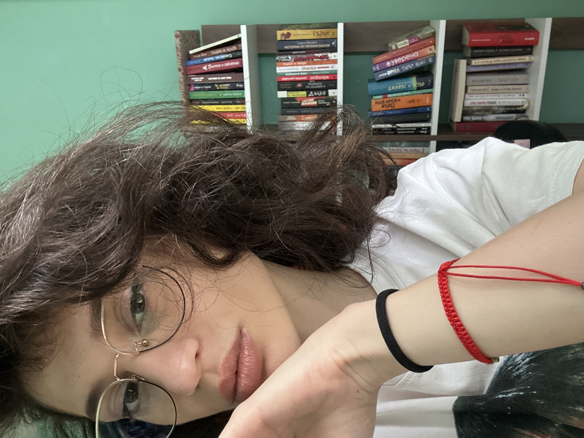 The image shows a close-up of a young woman with dark, wavy hair and glasses. She is wearing a white t-shirt and has a red bracelet on her wrist. She is resting her chin on her hand, which is also adorned with a black bracelet. In the background, a bookshelf filled with books is visible against a teal-colored wall. The environment suggests an indoor setting, possibly a home library or study. The specific location in the city of Sofia, Bulgaria cannot be determined from the visual information provided.