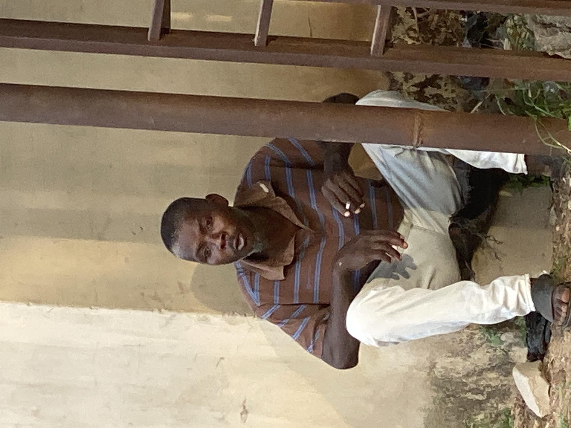 A man sits on the ground beneath a metal railing structure in Adankolo, Nigeria. He wears a brown striped shirt, white pants, and sandals. He holds something in his hand and looks at the camera. The area around him is a mix of bare ground and sparse vegetation. A concrete wall is visible in the background.