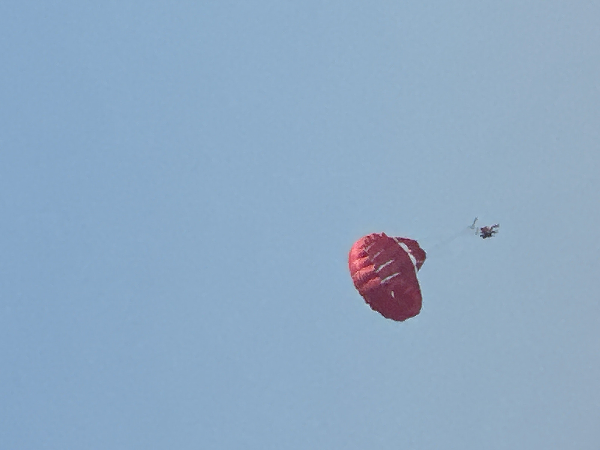 A single red parachute is visible against a clear blue sky. Two people are attached to the parachute harness, one above the other, appearing to be tandem parasailing. The parachute is situated in the upper left portion of the image, with the two individuals visible below it towards the bottom right of the parachute. The background is entirely a uniform light blue sky, with no other objects or discernible features. The image does not provide any information or context related to the city of Boreti, Montenegro.