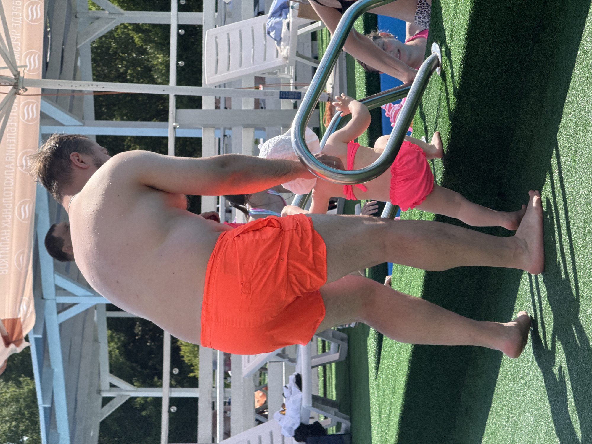 The image shows a family at a poolside in Koloksha, Russia. A man in orange swim trunks is in the foreground, his back to the camera. A young child in a pink bikini is climbing a pool ladder, being assisted by an adult whose arm is visible. Another child in a white hat and pink bikini bottom is near the ladder, and partially visible is another adult in a leopard print swimsuit. The setting appears to be a sunny day at an outdoor pool, with green artificial turf surrounding the water. White lounge chairs and a shaded area with a banner are visible in the background. The banner has text which appears to be in Cyrillic. The overall mood seems relaxed and family-oriented, enjoying a day at the pool.