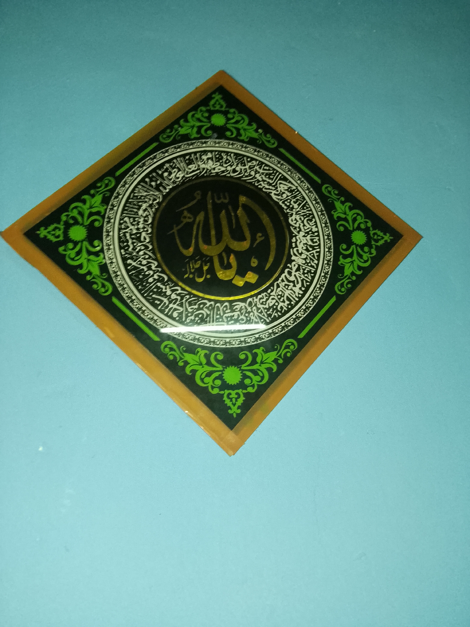 A diamond-shaped Islamic calligraphy artwork is displayed on a light blue wall. The artwork features a central circular design with Arabic script in gold, set against a black background. This central design is surrounded by intricate white Arabic calligraphy. The entire circular motif is enclosed within a green border adorned with floral patterns. The artwork itself is framed by a dark green band, and the outermost edge is a golden-brown border. The image is taken from a slightly elevated angle, looking down at the artwork. The lighting appears to be artificial, casting a slight sheen on the artwork. The background is a plain, textured light blue wall. This artwork is likely a decorative piece hanging in a residential or public space in the city of Bencongan, Indonesia.