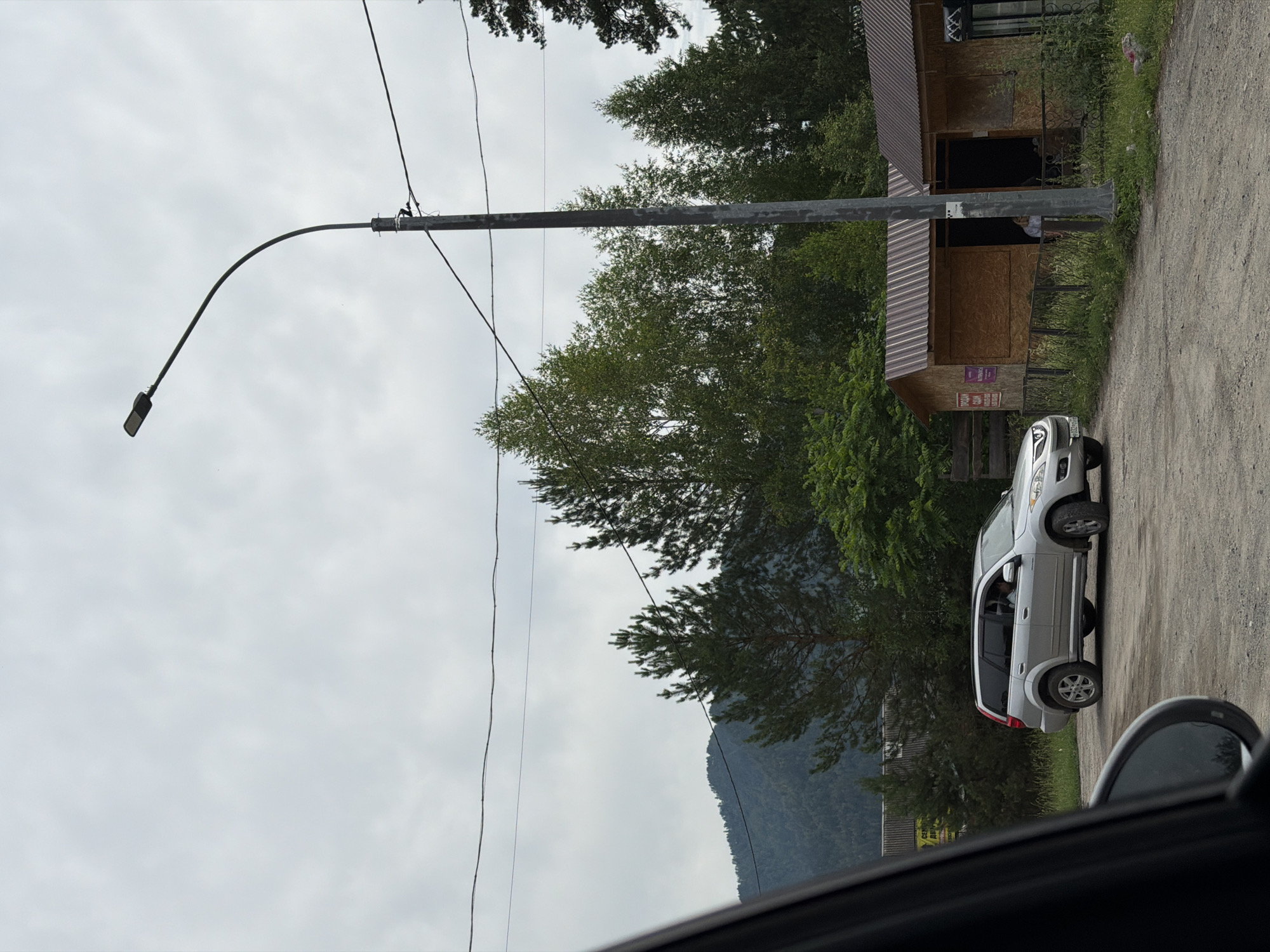 A silver SUV is parked on a dirt road next to a small wooden building with a corrugated metal roof. A person is visible inside the building. The scene is outdoors, with trees and a mountain in the background. A street lamp stands on the right side of the image. The sky is overcast. This appears to be the village of Ust-Sema, Russia, given the context.