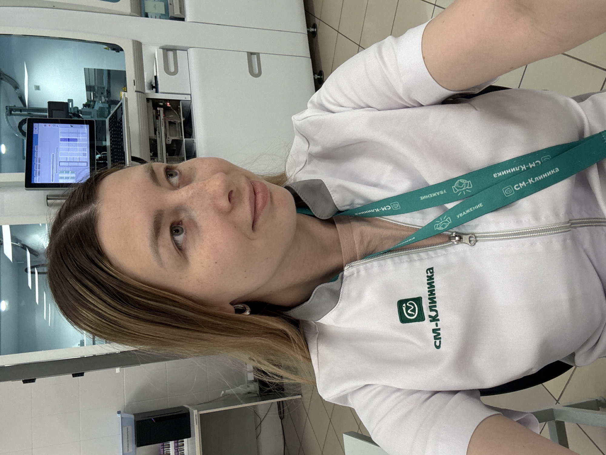 A young woman is pictured in what appears to be a medical laboratory setting. She is wearing a white lab coat with the inscription "CM-Клиника" (SM-Clinic) and a green lanyard around her neck with the same logo and Russian words that translate to "Respect." The background reveals sophisticated lab equipment, indicating an advanced medical environment. The location is likely within a "SM-Clinic" facility in Moscow, Russia, as indicated by the Cyrillic text on the coat and lanyard.