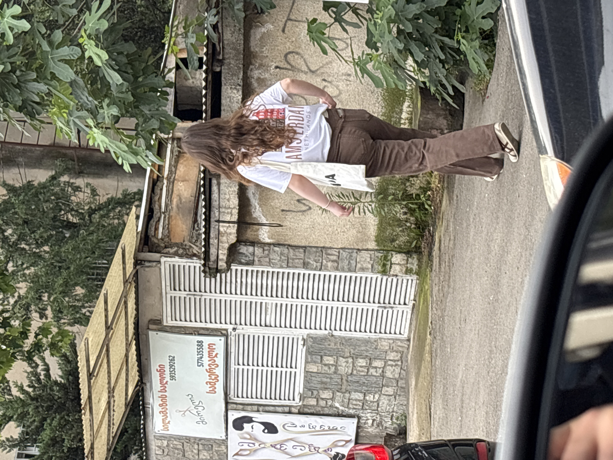 The image shows a street scene in Tbilisi, Georgia. A young woman with long, wavy brown hair is walking away from the camera, wearing a white t-shirt with "AMSTERDAM NETHERLANDS" printed on the back and brown pants. She is carrying a white tote bag. Behind her is a building with a corrugated metal awning over a sign written in Georgian script, which appears to be for a barbershop named "Anareli" with phone numbers 593529262 and 577435588. The building is constructed of stone and has white louvered shutters on a window and door. The street is paved, and there is greenery and trees in the background. A car is partially visible in the foreground, suggesting the photo was taken from another vehicle.