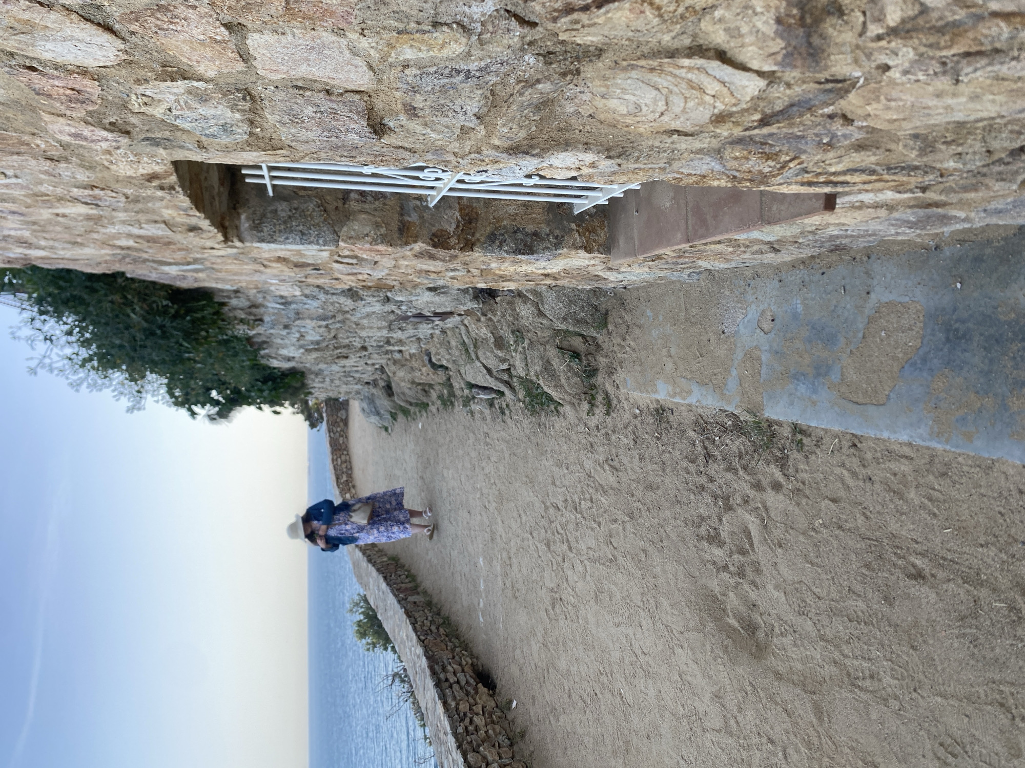 A person is standing on a path alongside the sea in Palafrugell, Spain. The person is wearing a hat, a blue floral dress, and a denim jacket. The path is bordered by a low stone wall on one side and a rough stone structure above. A glimpse of the blue sea and sky is visible. The scene is taken from a slightly elevated perspective, looking down the path.