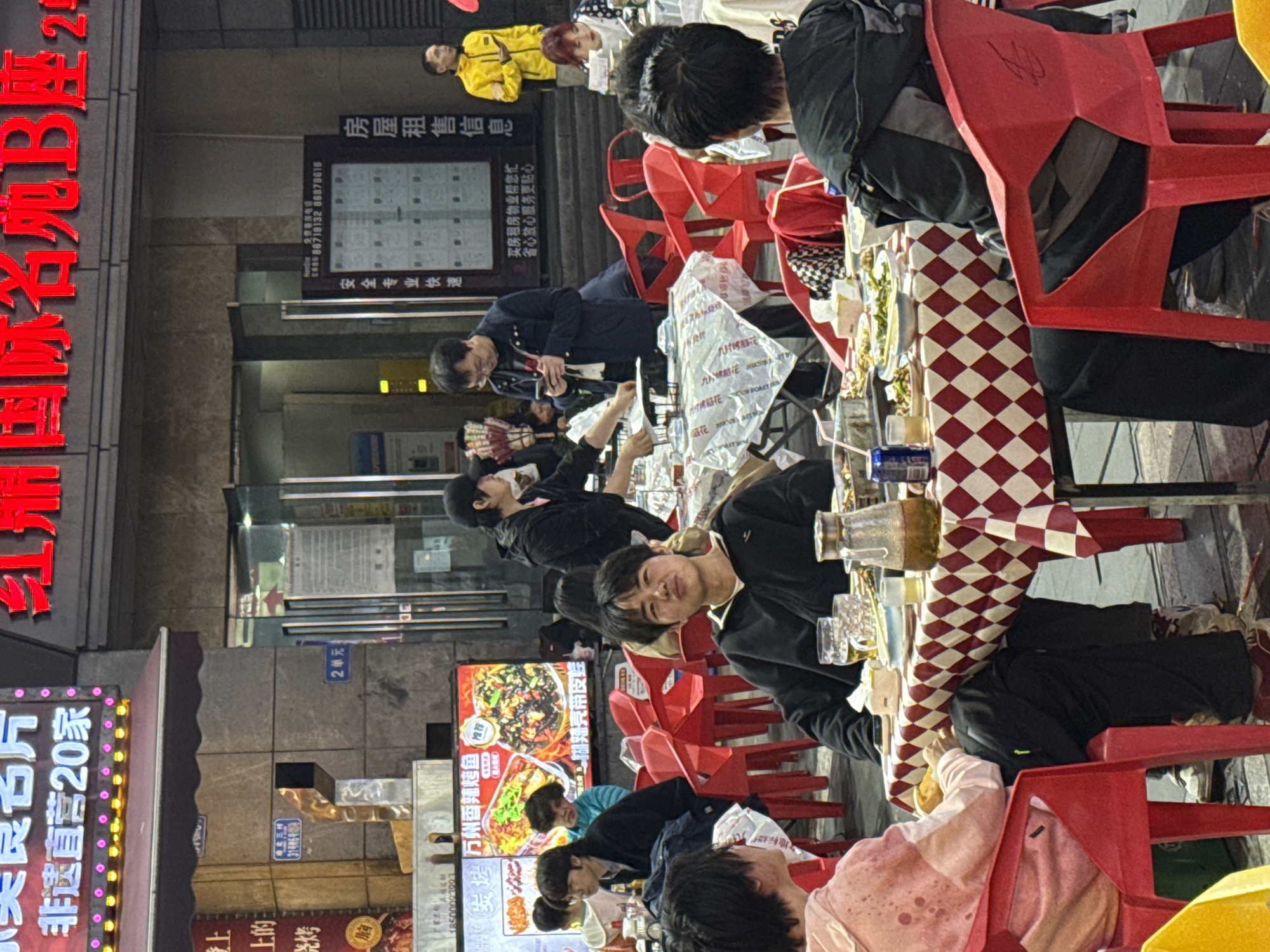 An outdoor evening scene depicts a bustling street dining area in Liangjiang, China. Multiple patrons are seated at square tables draped with red and white checkered tablecloths and furnished with red plastic chairs.
In the mid-foreground, a young male wearing a black zippered hoodie is seated, facing the viewer with a slight upward gaze and pursed lips. On his table, a large glass mug filled with a pale liquid, a pitcher containing a dark liquid, a blue beverage can, and several dishes with food remnants are visible. To his left, another individual in a pink and white tie-dye hoodie is partially visible, facing away. To his right, a person in a dark jacket is also seated, facing away from the camera.
In the mid-background, multiple individuals are engaged in dining or activity. A person in a black cap and jacket appears to be attending to a table covered with a white plastic sheet. Nearby, a man in a dark blue jacket and glasses stands, looking down at a smartphone. Further back, a man in a bright yellow jacket stands, similarly engaged with a smartphone.
The background consists of commercial buildings illuminated by vibrant neon signs featuring Chinese characters. A prominent red sign reads "红鼎国际名苑B座" (Hongding International Famous Garden Block B), and a pink and white sign above it states "美味名片 非达直营20家" (Delicious Business Card, 20 direct stores). Large illuminated display screens show images of food, including "万州香辣烤鱼" (Wanzhou Spicy Grilled Fish). A vertical sign advertises "房屋租售信息" (Housing Rental Information) and "安全专业快速" (Safe, Professional, Fast) with associated contact numbers. The ground appears to be paved, and the overall lighting suggests evening or nighttime.
