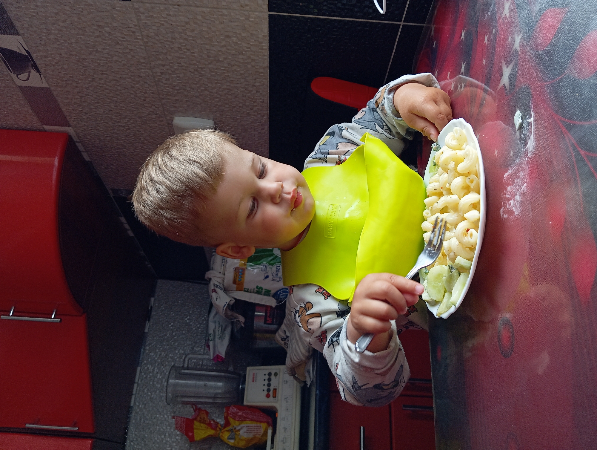 A young boy with blond hair is sitting at a table, eating pasta with a fork. He is wearing a yellow bib and a long-sleeved shirt with a cartoon pattern. The table has a red and black patterned surface. In the background, there are red kitchen cabinets, a blender, and various food items. The scene appears to be in a kitchen. The location is Hlynytsi, Ukraine.