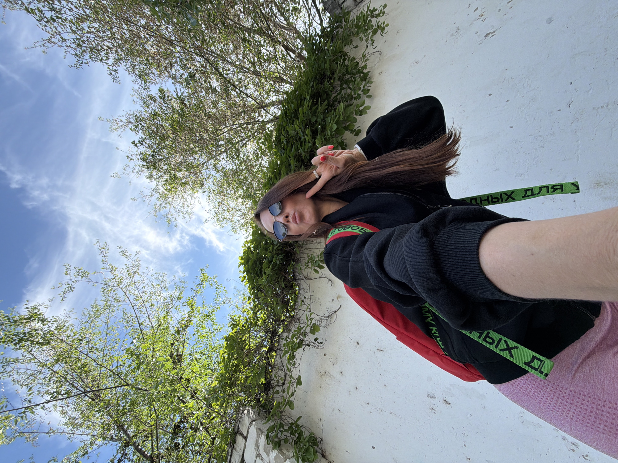 A low-angle, wide shot shows a young woman with long brown hair wearing sunglasses, a black zip-up hoodie, and pink pants. She is holding her right hand up with two fingers extended in a peace sign and blowing a kiss. A red backpack is visible over her left shoulder. She is standing in front of a white textured wall covered with ivy, with green trees and a blue sky with wispy clouds visible above. A green strap with black lettering is visible on her hoodie. The image is taken from the perspective of someone holding a camera or phone, as her arm is extended. The scene appears to be outdoors in an urban or semi-urban environment.