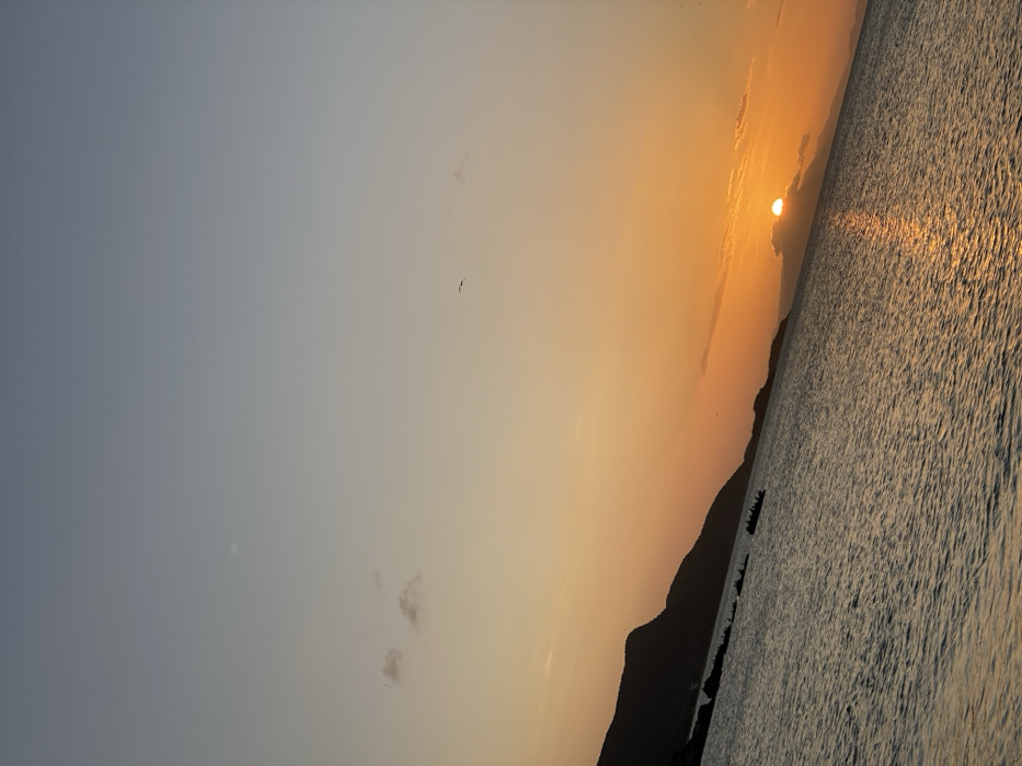 A sunset view with a hazy sky transitioning from light blue to orange. The sun is partially obscured by clouds, casting a golden reflection on the water. A silhouette of a mountain range lines the water's edge. A bird is visible in the sky.